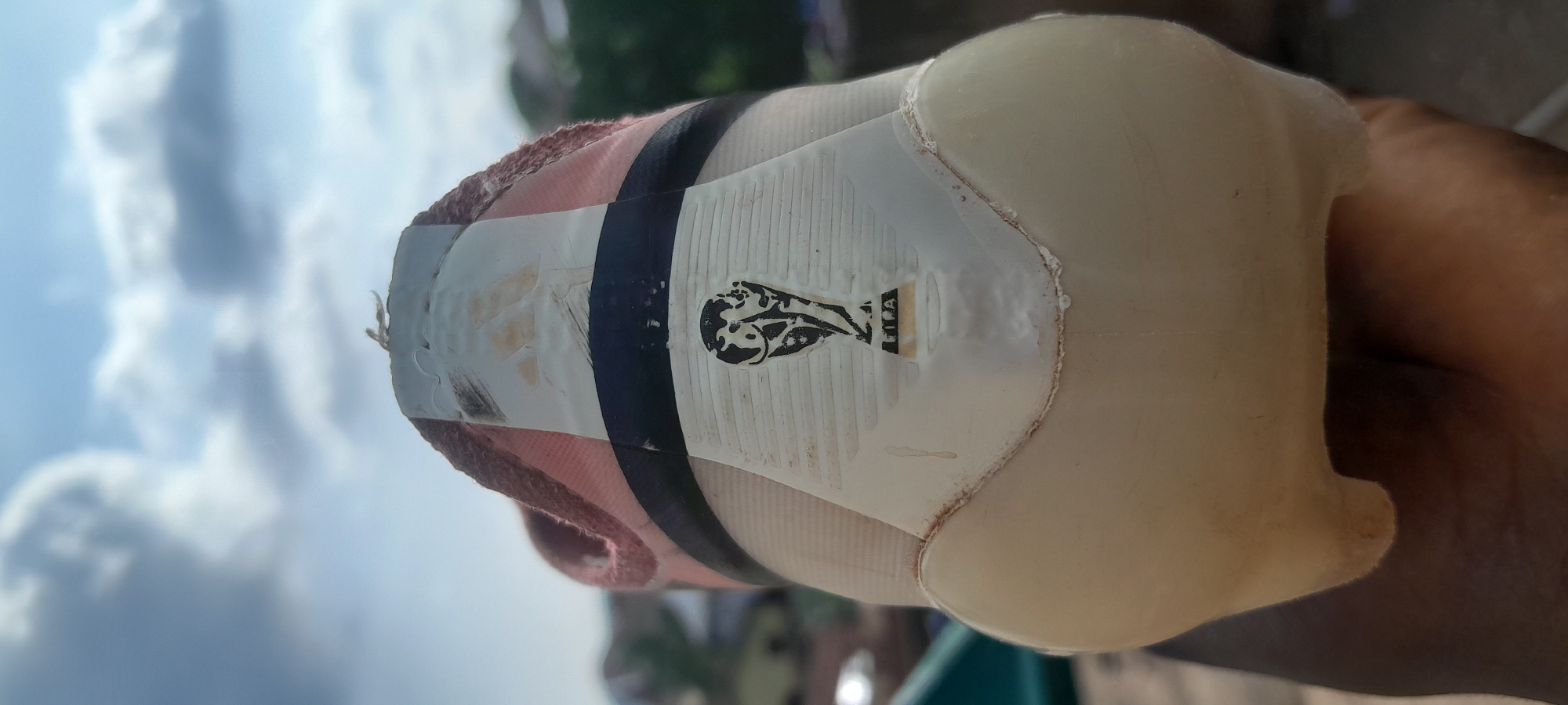 A close-up, low-angle shot focuses on the heel of a pink and white soccer cleat, held in a dark-skinned hand, set against a bright, cloudy sky. The cleat features a black stripe across the heel and a faded FIFA World Cup trophy logo. The shoe appears worn and slightly dirty. In the background, blurred buildings and greenery suggest an outdoor urban setting in Abuja, Nigeria. The lighting indicates it is daytime.
