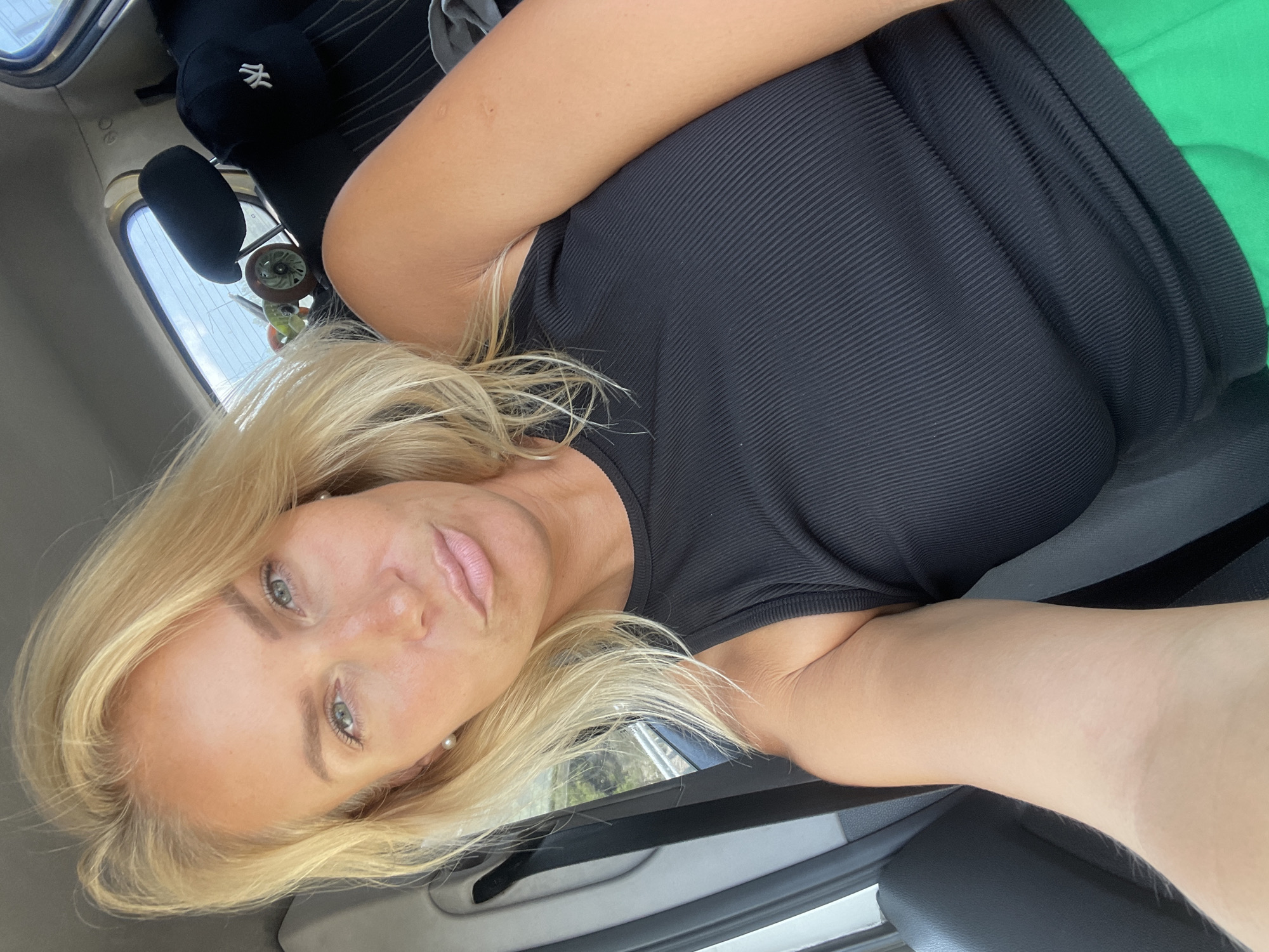 A blonde woman is taking a selfie in a car. She is wearing a black ribbed tank top and green pants. Her hair is blonde and wavy. She has green eyes and light skin with freckles. She is wearing pearl earrings. The car interior is visible, including the headrest and a black baseball cap with a white "NY" logo. Outside the window, there is greenery and a body of water, suggesting a coastal location. The image appears to be taken during the daytime. The setting is consistent with Gelendzhik, Russia, known for its coastline and greenery.