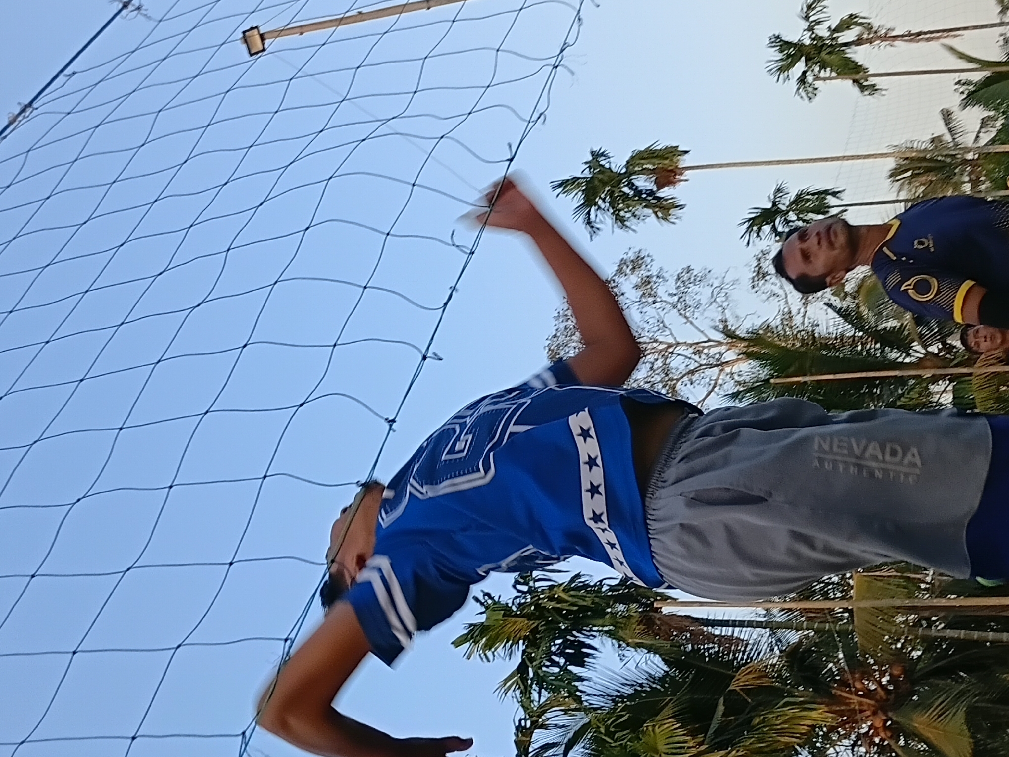 In Margo Rukun, Indonesia, two men are engaged in a volleyball game, with one player in the foreground executing a jump serve over a net. He is wearing a blue jersey with white accents and gray athletic shorts. A second man, positioned further back, observes the game. The background features a clear sky, palm trees, and a tall pole with a light fixture. The overall scene captures a moment of athletic activity in an outdoor, tropical setting.
