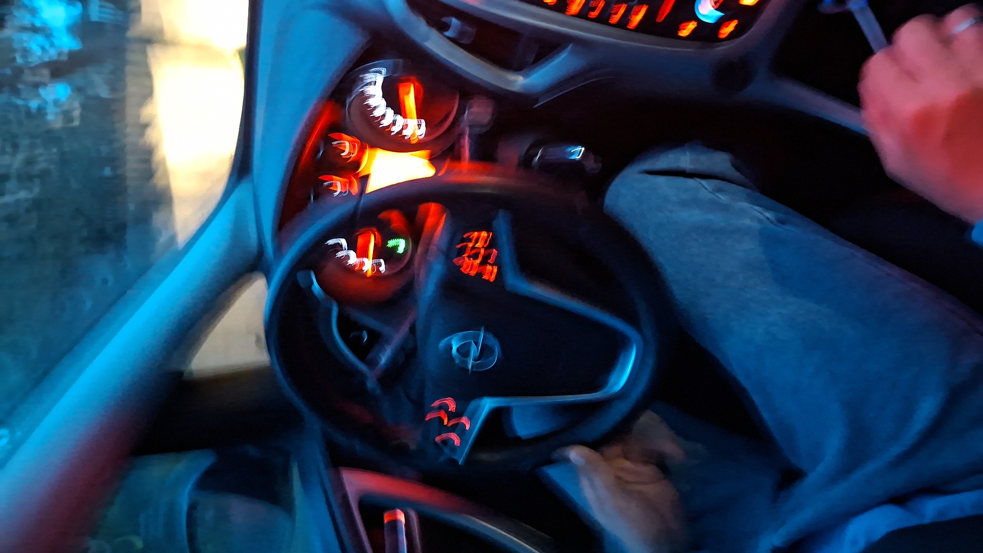 Interior view of a car, likely in motion due to motion blur. The steering wheel is visible with a logo on it. Car dashboard gauges are lit in red. Person's hands are partially visible on the steering wheel. A leg wearing denim jeans is also in the frame. Out the front windshield can be seen a blurry mix of yellow and blue lights. The image provides no indication of the location being Забалоцкі сельскі Савет, Belarus.