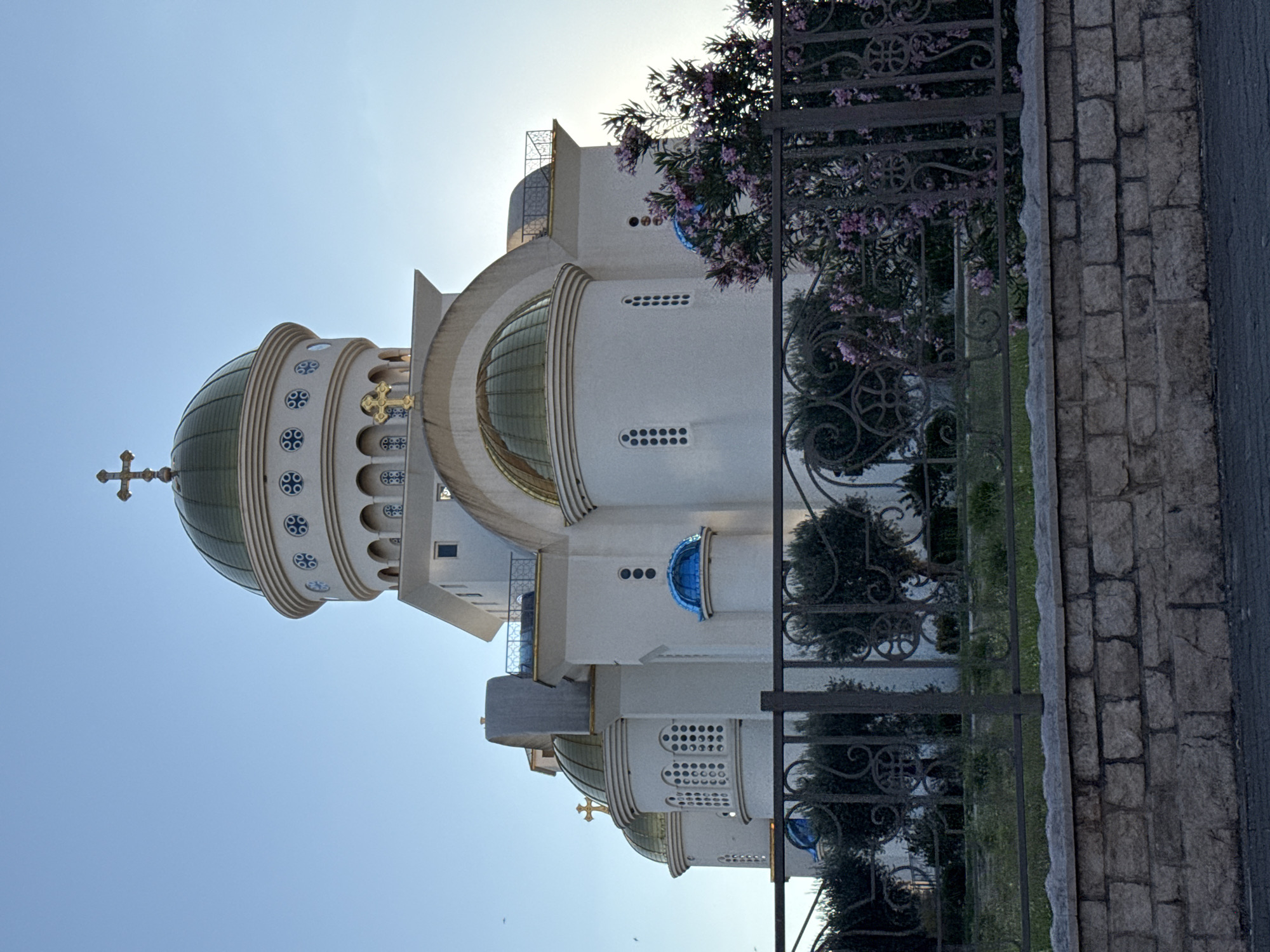 The image shows the Church of St. Jovan Vladimir in Bar, Montenegro, a prominent religious building characterized by its white facade and golden domes. The church features multiple towers topped with crosses, and its architectural style includes arched windows and decorative elements. In the foreground, a metal fence with ornate scrollwork separates the viewer from the church grounds, which include green bushes and flowering plants with pink blossoms. A stone wall runs along the bottom of the image, leading to a paved area. The sky is clear and blue, indicating daylight, likely late afternoon given the lighting. There are no people visible in the frame.