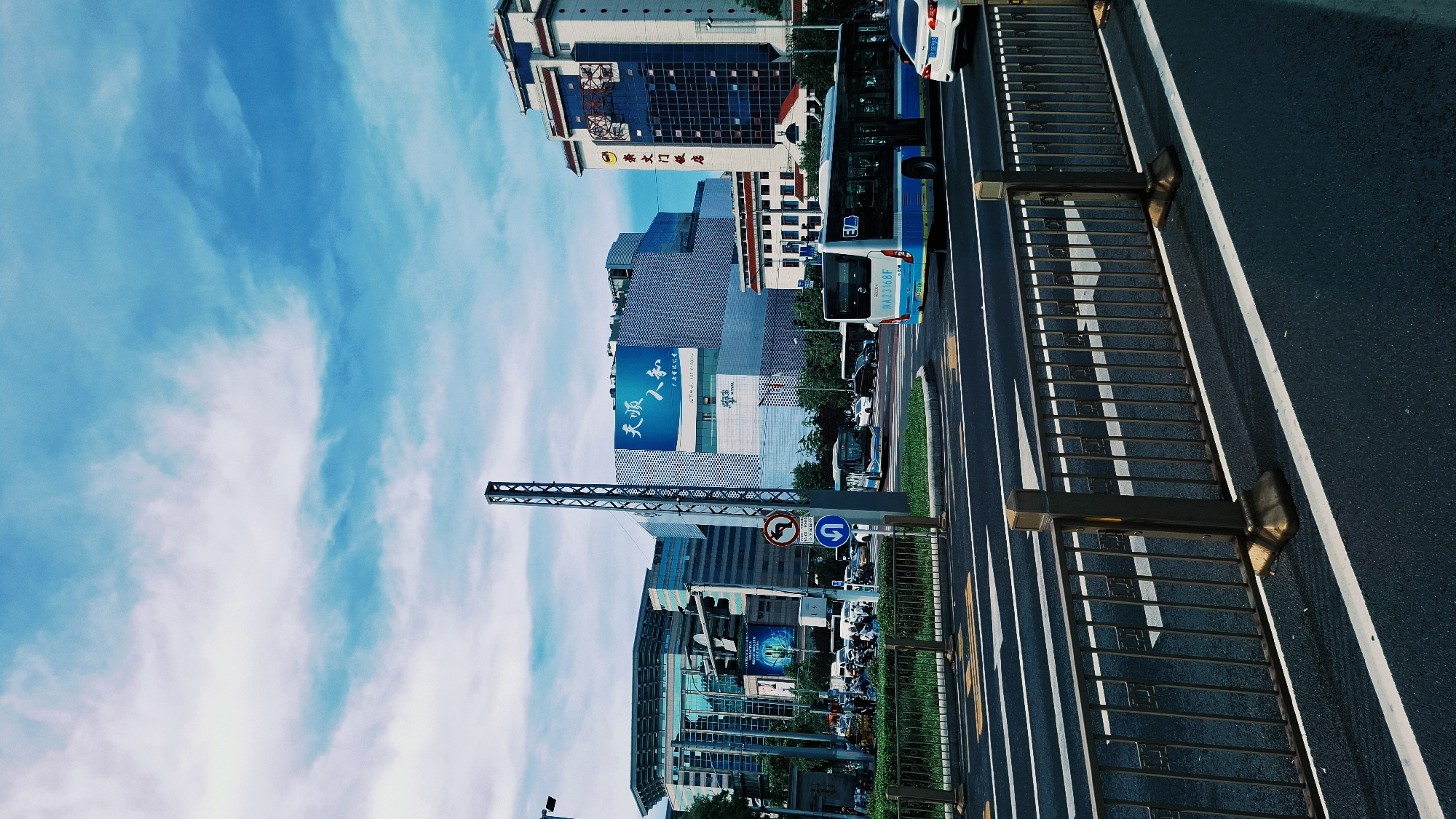 The image captures a street scene in Dongcheng District, China. The perspective is tilted 90 degrees clockwise. Buildings line the street, some displaying Chinese characters on their facades. A blue bus is visible on the road. A pedestrian walkway with metal railings runs along the side of the road. Overhead, a sign structure spans the road. The sky is visible behind the buildings, with scattered clouds.