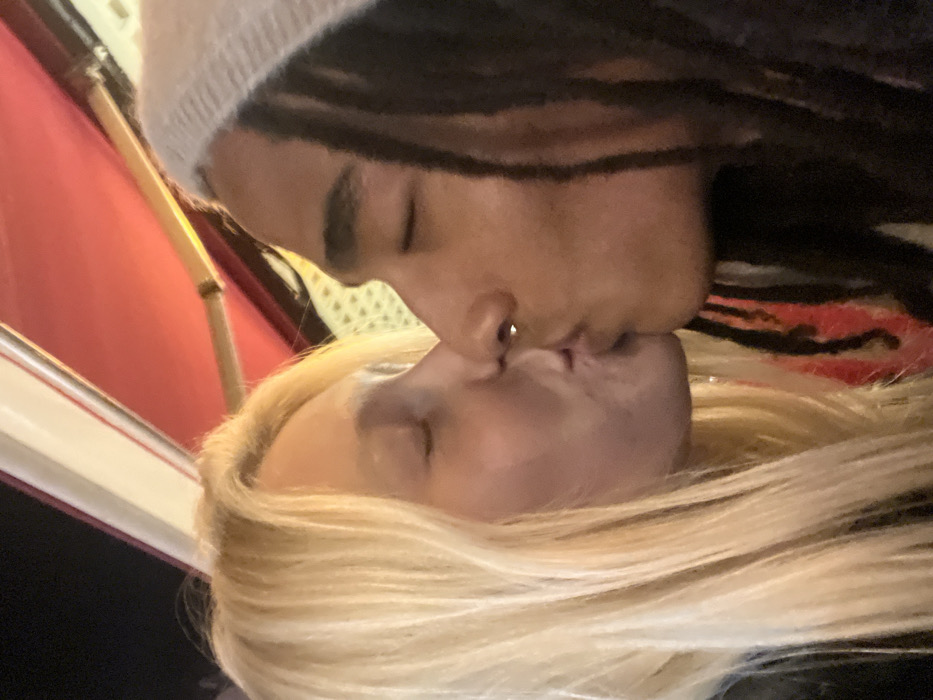 Two individuals are closely embraced, with their lips meeting in a kiss. One person has long, blond hair, while the other has dreadlocks and wears a beige beanie. The photo appears to be taken indoors, with glimpses of red and off-white elements in the background. The lighting is natural.
