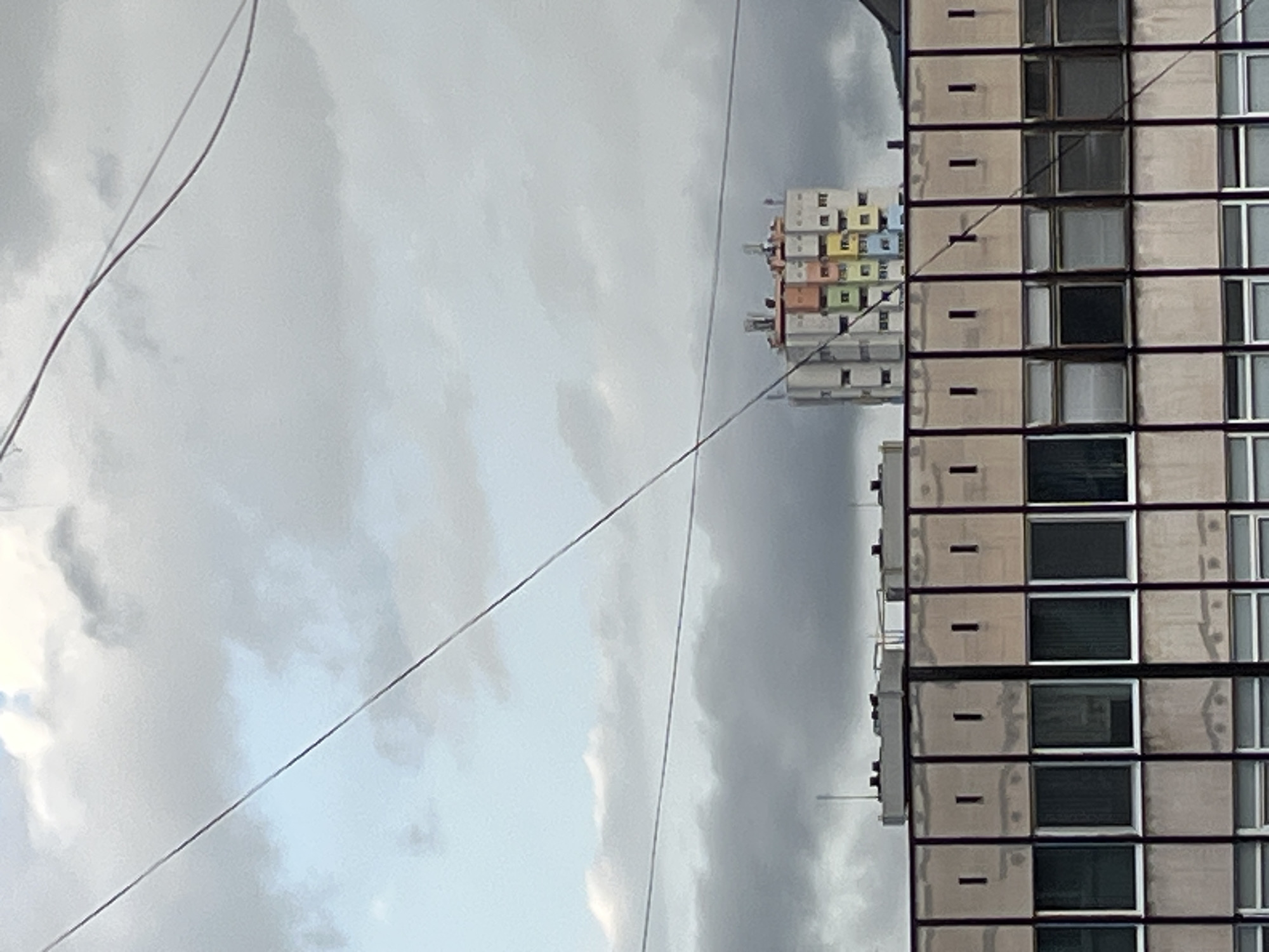 This media file captures an outdoor urban or suburban scene, rotated 90 degrees to the left, so that buildings appear to be lying horizontally against a cloudy sky.
On the right side of the frame, a large, multi-story building dominates, stretched horizontally across the image. It features a regular grid pattern of windows and beige-colored panels, suggesting a modern architectural style, possibly residential or commercial. The windows are dark, some reflecting light, and some appear to have blinds. Small, dark, horizontal details are visible on the beige panels between floors.
Further to the left and in the distance, another, smaller building is visible. This structure is composed of numerous stacked, box-like units, each appearing to be painted a different pastel color, including yellow, green, blue, orange, and pink. This building also appears horizontal in the current orientation.
The majority of the background and foreground is filled with a cloudy, overcast sky. The clouds are a mix of light grey and white, indicating an overcast daytime. Several thin, dark wires or cables crisscross the sky diagonally, extending from the upper left towards the bottom middle of the frame, passing between the viewer and the distant buildings. One thicker wire is also visible in the upper left.
There are no people or specific activities depicted in the image. The scene is static. There is no visible text. The weather appears to be a cloudy day.