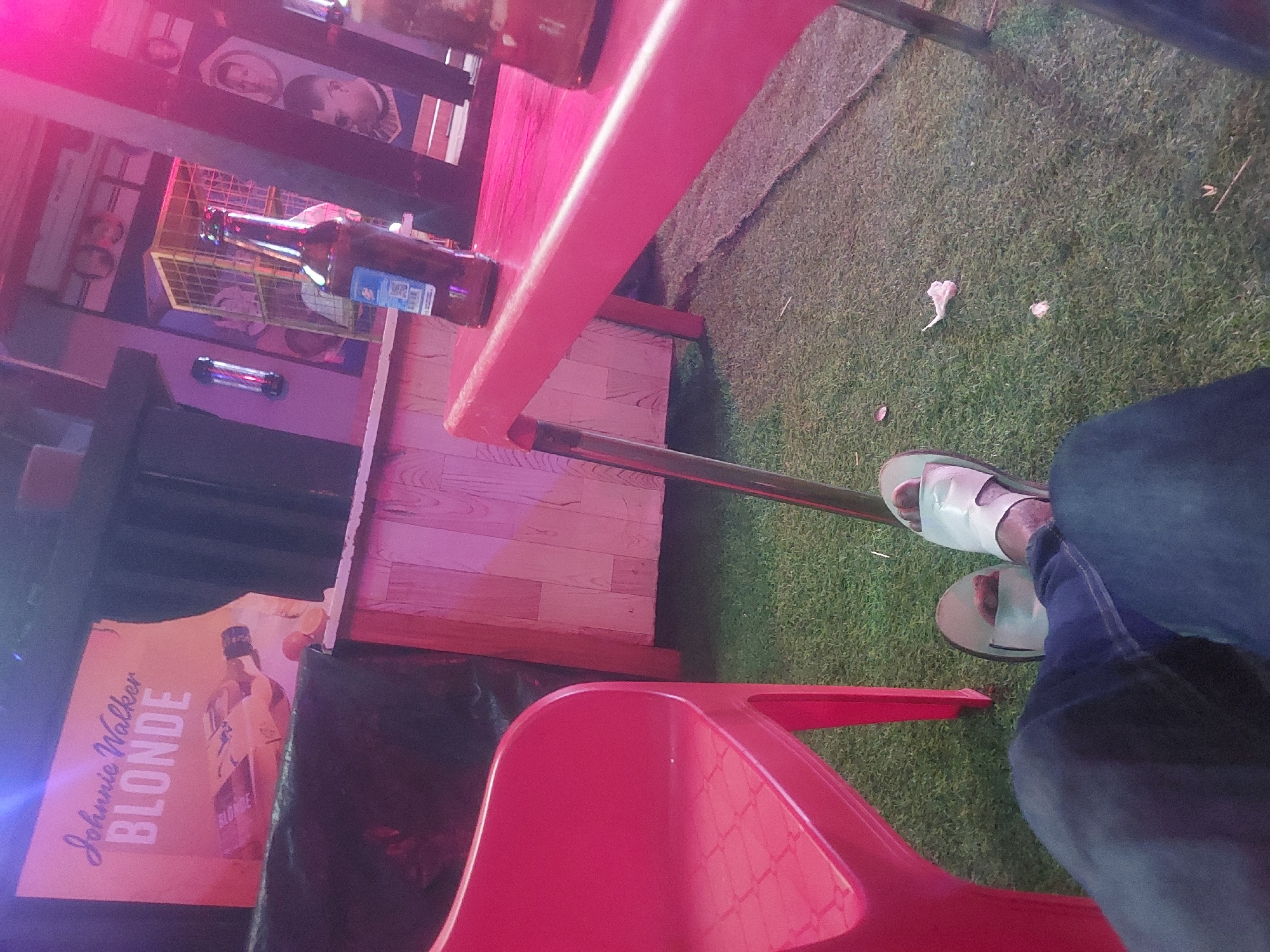 The scene appears to be a casual outdoor bar or restaurant setting, possibly in Dar es-Salaam, Tanzania, as inferred from the environment and beer bottle.
Environment: The setting includes artificial turf flooring. The lighting is predominantly pink/red, suggesting artificial or neon lighting.
Objects: A red plastic chair is visible, along with a pink table where a beer bottle rests. A "Johnnie Walker Blonde" promotional poster is present. Debris, possibly paper scraps, are on the turf.
People: The image includes a partial view of a person wearing blue jeans and light-colored sandals.
Actions and Interactions: The person is seated, possibly at the table. A beer bottle is on the table, suggesting a social gathering or relaxation.
City Context: Dar es-Salaam is inferred as a possible location, though not explicitly depicted.
