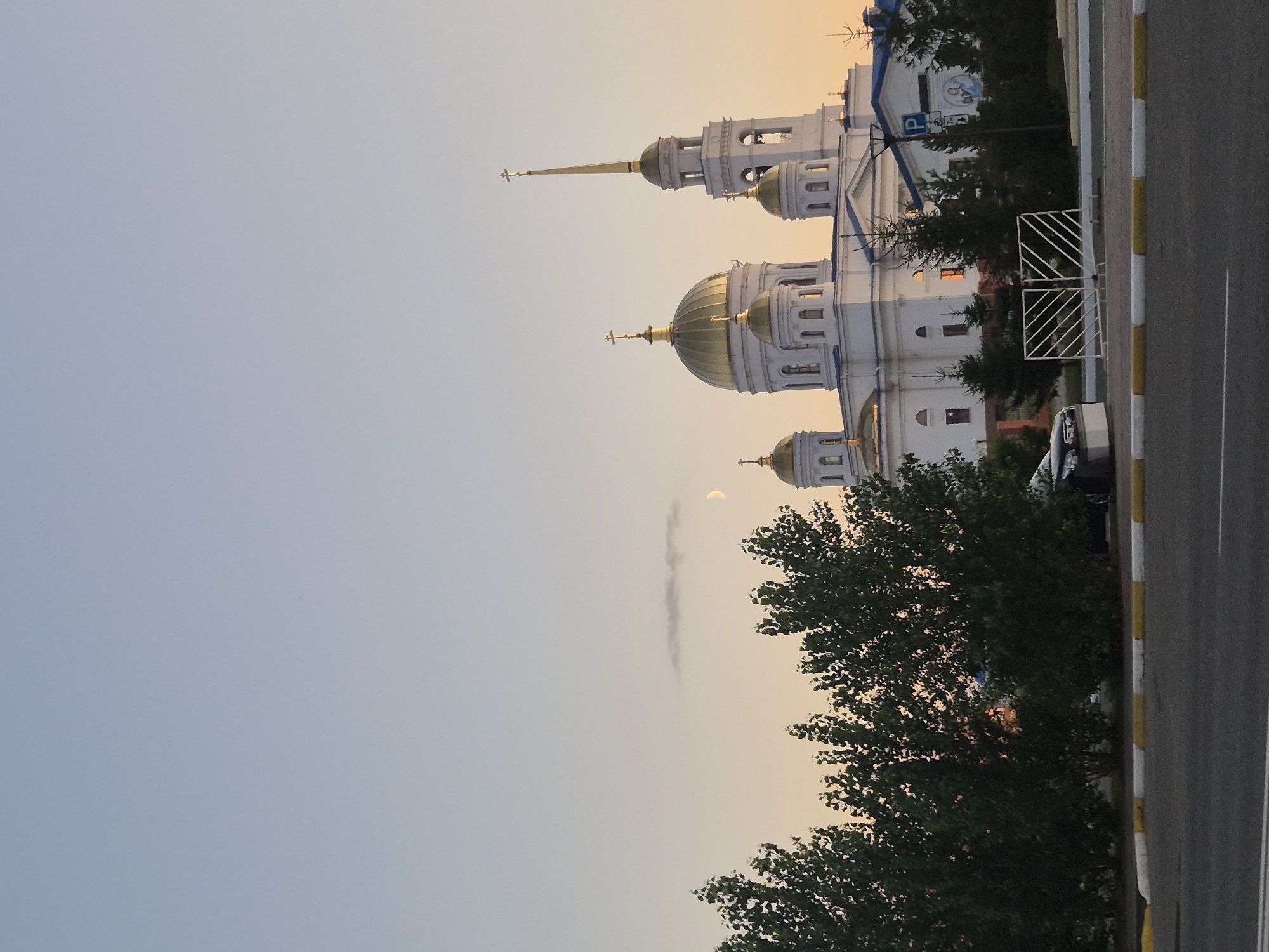 The image shows a large white cathedral with golden domes in Kokshetau, Kazakhstan. The cathedral is situated behind a row of green trees and a white metal fence. A crescent moon is visible in the pale sky, along with a wispy cloud. In the foreground, there is a road with a yellow and white striped curb. A dark-colored car is parked near the fence, and a blue parking sign with a white "P" is mounted on a pole to the right of the cathedral. The overall scene suggests a serene evening or early morning atmosphere.