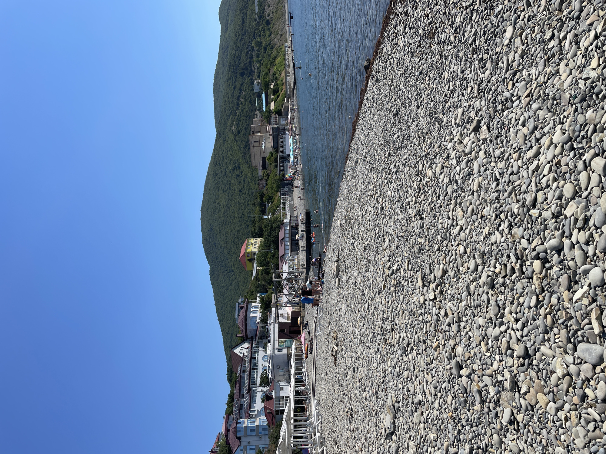 The image depicts a pebble beach in Shirokaya Balka, Russia, on a clear, sunny day. The foreground is dominated by a dense scattering of smooth, grey and brown pebbles. In the midground, a body of calm, blue water laps the shore. Several people are visible along the waterline and in the sea. Some are standing or walking on the beach, while others are swimming or engaged in water activities. Beach umbrellas and lounge chairs are set up on the pebbled area. Behind the beach, a stretch of buildings, including multi-story structures with red-tiled roofs and white facades, line the coastline. Further inland, a lush green, tree-covered hillside rises steeply, creating a scenic backdrop. The sky is a vibrant, cloudless blue.