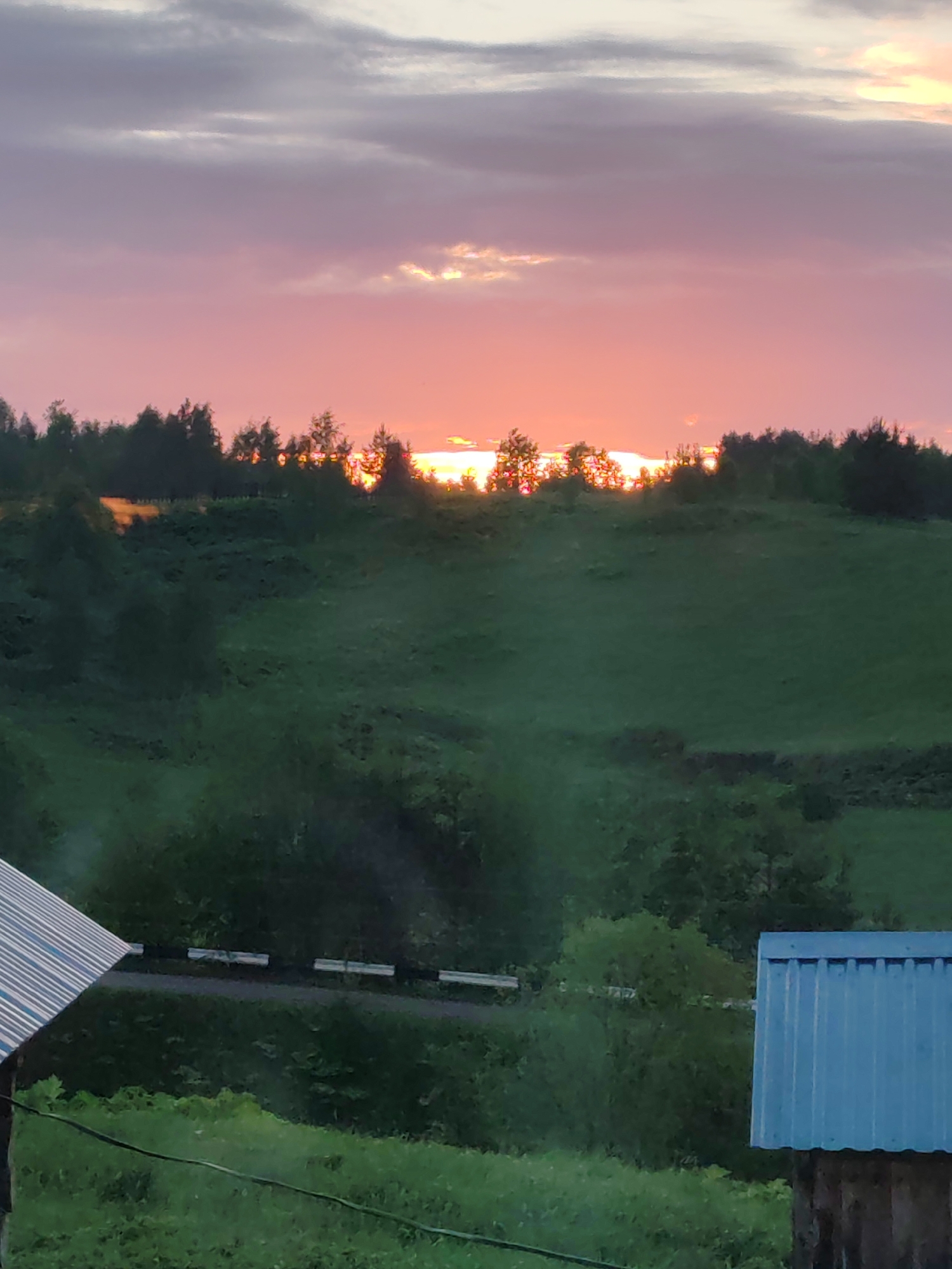 The photograph depicts a sunset in a rural area, possibly near or in Чухлэм, Russia. The sky is predominantly pink and purple, with a sliver of bright orange light visible on the horizon where the sun is setting behind a line of trees. The foreground features green hills and foliage, indicating a natural or agricultural landscape. Parts of two buildings are visible, suggesting the presence of human habitation. A road runs horizontally across the image.