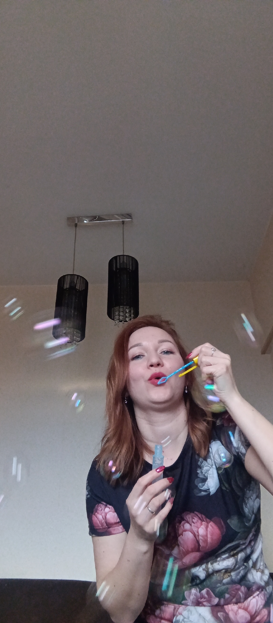 A woman with red hair and red lipstick is blowing bubbles indoors. She is wearing a floral shirt and a wedding ring on her left hand. She holds a blue bottle of bubble solution in one hand and a yellow and blue bubble wand in the other. Bubbles are visible around her. In the background, there is a light fixture with two black shades hanging from the ceiling, which appears to be white. The walls are light beige.