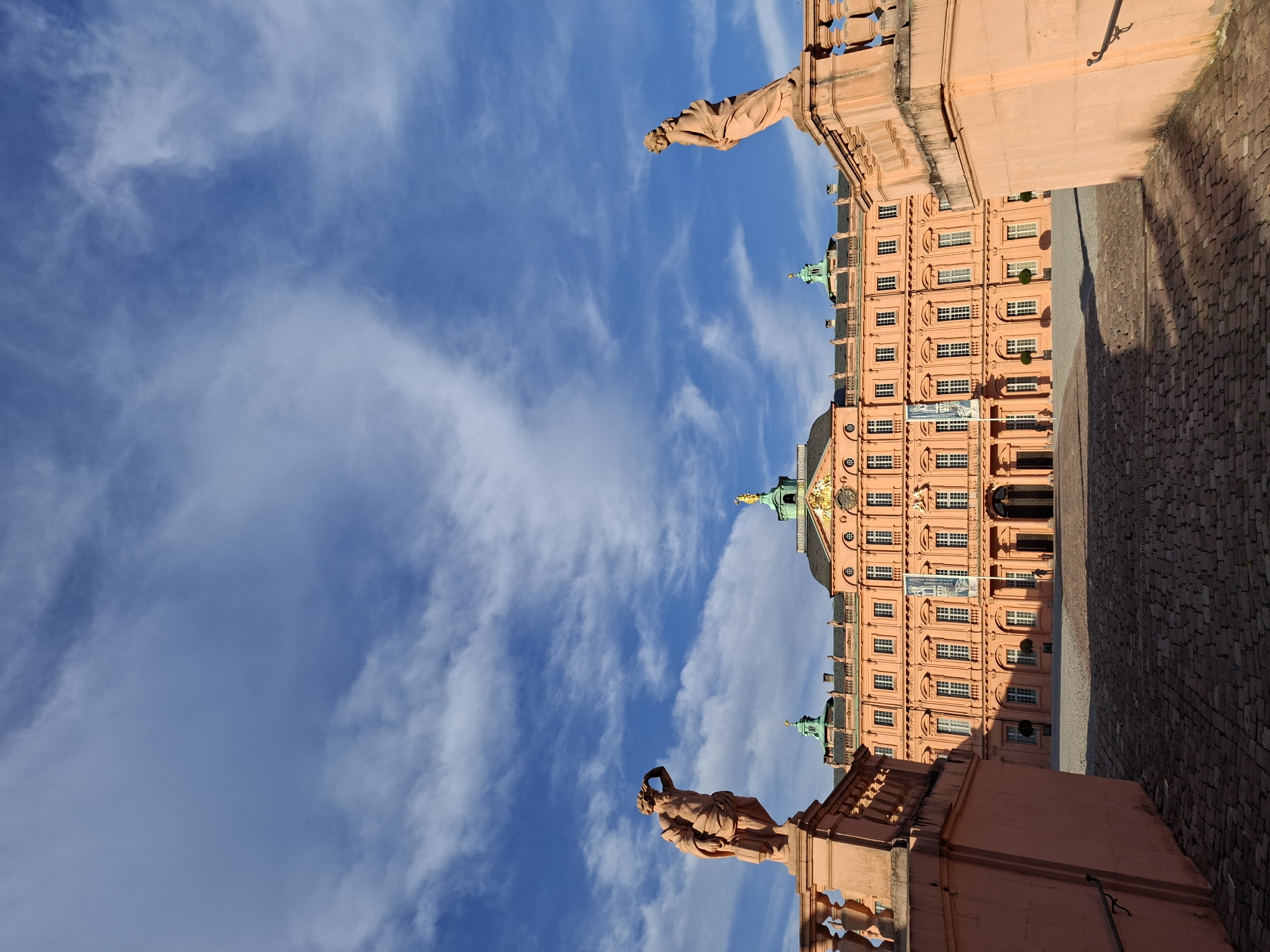 The image shows the Rastatt Residential Palace in Rastatt, Germany. The palace is a large, pink Baroque building with a green-domed central tower. The foreground features a cobblestone courtyard and two ornate stone balustrades topped with statues. The sky above is blue with scattered white clouds.