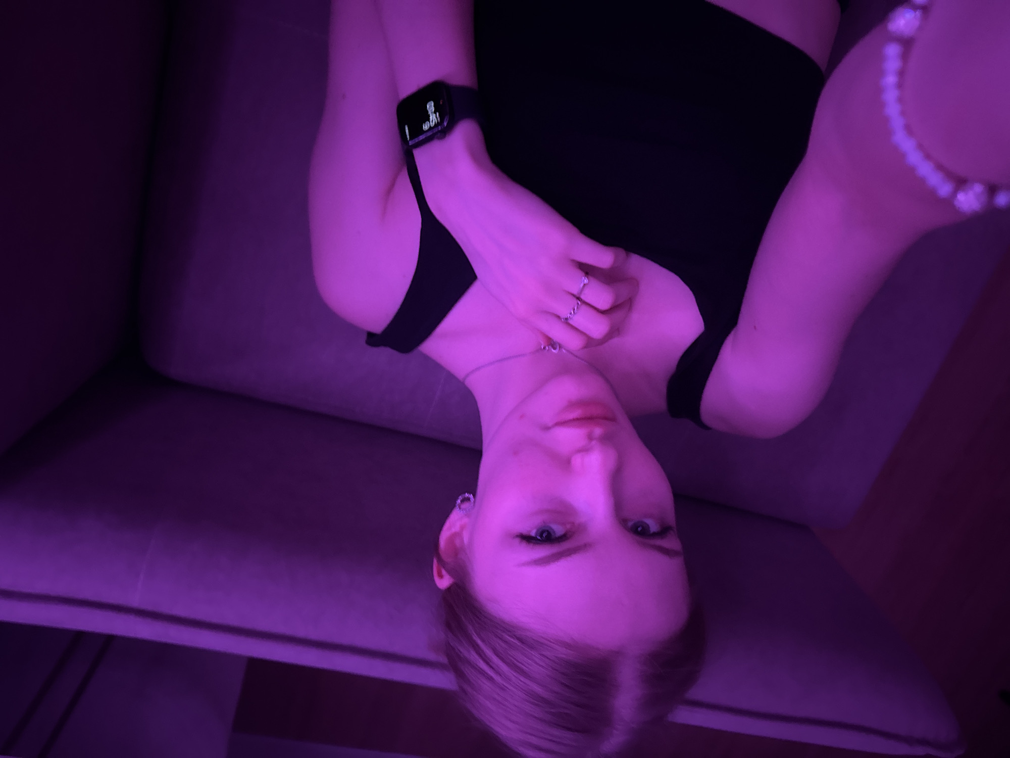A woman with pale skin and blonde hair, wearing a black top, lies on a gray couch. She is angled in the frame with her head near the bottom and torso extending upwards. A watch is visible on her left wrist and jewelry on her right wrist. A ring adorns one of her fingers. The scene is lit with a purple hue. The location of Perm, Russia cannot be determined from the visual information provided.