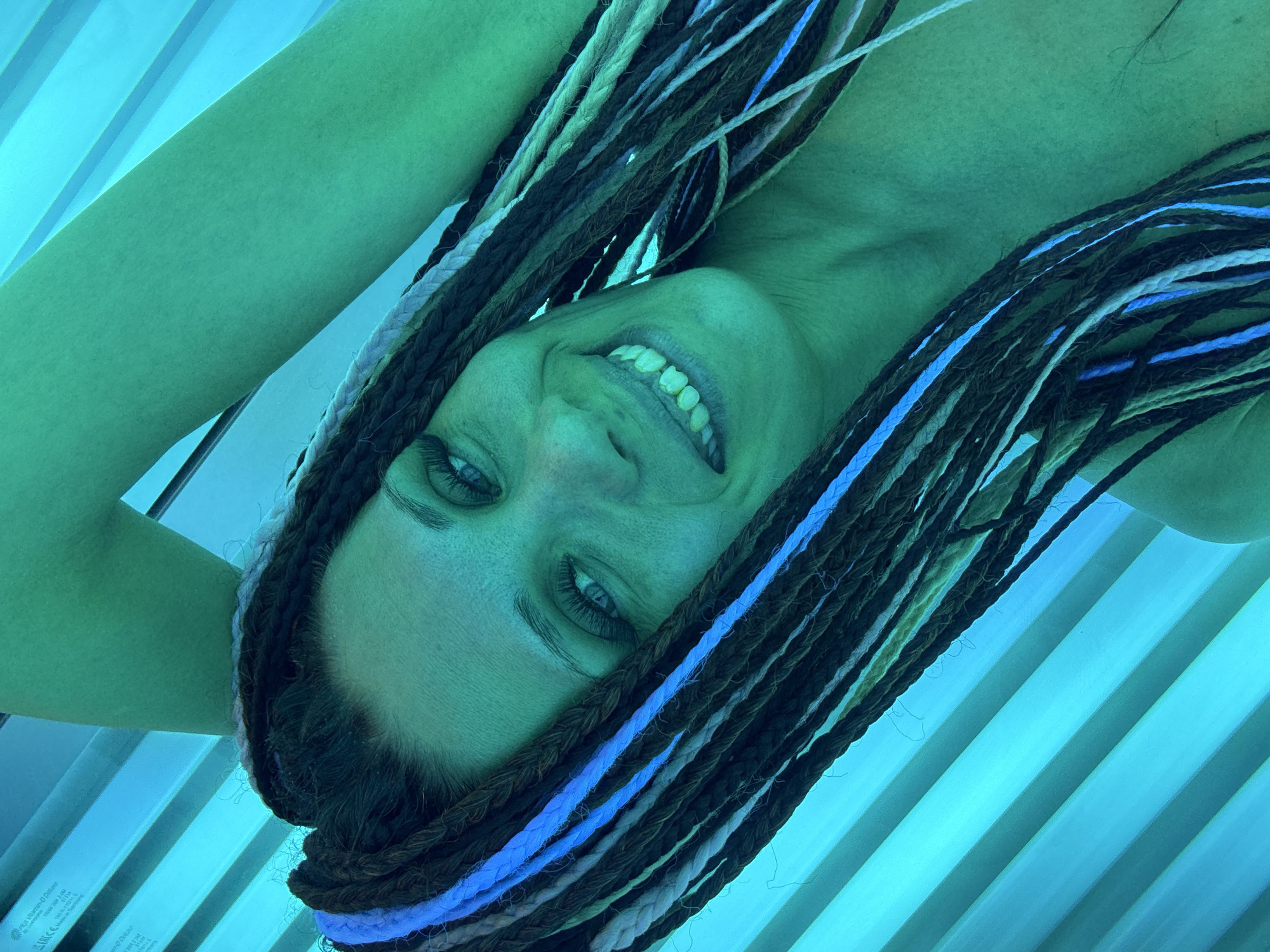 A woman with tan skin and braided hair lies inside a tanning bed in an unspecified location in the city of Unknown, Unknown. She smiles broadly, exposing her teeth. Her left arm is raised above her head. The tanning bed is illuminated with a blue-green light.
