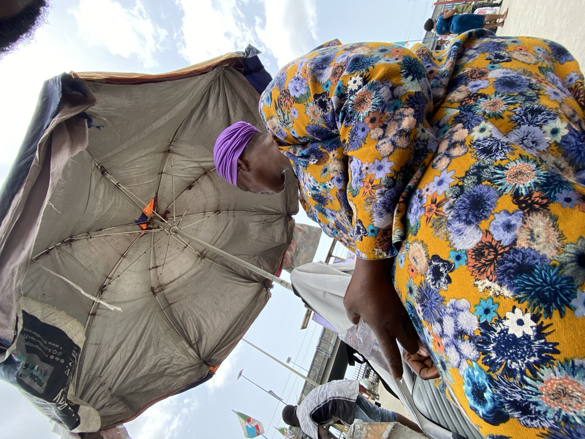Here's a detailed description of the image:
The photograph appears to be taken from a low angle, looking up at a scene that includes a woman in a floral patterned dress and a makeshift structure with a large, tattered umbrella. The woman is wearing a purple head covering and appears to be leaning over or looking down at something not visible in the frame. Her dress features a yellow background with a dense floral pattern of blues, purples, and oranges.
Overhead is a large umbrella-like structure, likely intended for shade. The umbrella's covering is worn and faded, appearing grey with visible stains and possibly made from repurposed materials. The metal ribs of the umbrella are visible.
In the blurred background, other people and structures are discernible. There is a pedestrian bridge or elevated walkway visible. Flags are seen lining the road.
Given that the image mentions Lekki, Nigeria, the scene likely portrays a street-side vendor or informal business operating under rudimentary shelter, a common sight in many parts of the city. The presence of flags and a pedestrian bridge suggests a busy area, potentially a market or roadside business district.