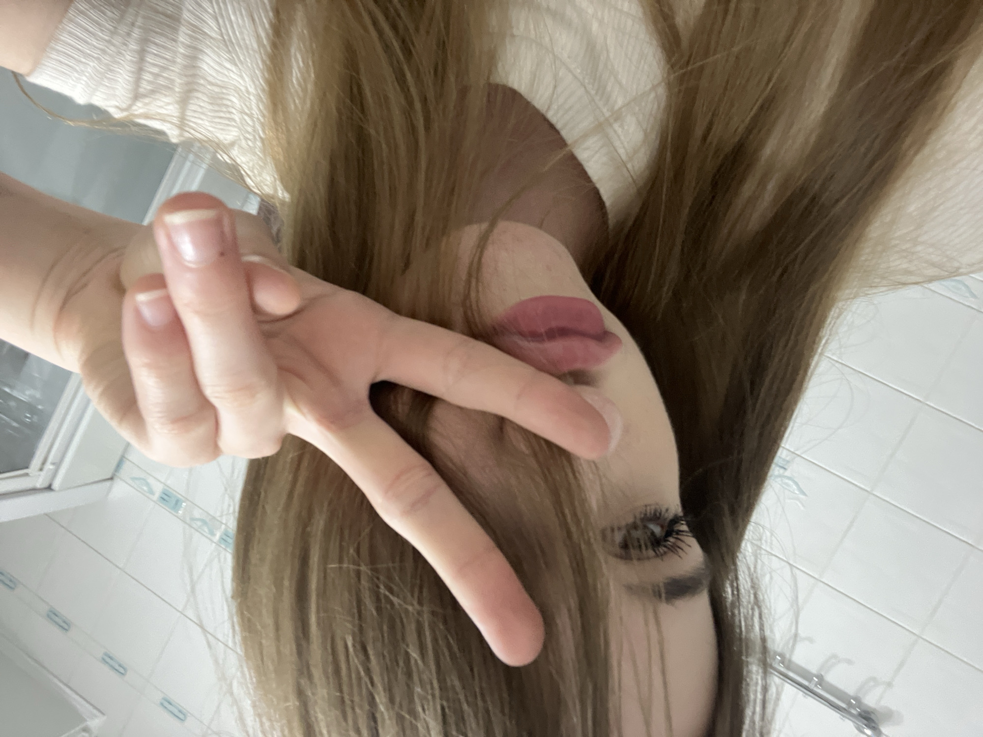 A young woman with long, brown hair and dramatic eye makeup is taking a selfie in what appears to be a bathroom. She is wearing a cream-colored, ribbed shirt. Her right hand is raised, with her index and middle fingers extended in a peace sign, partially obscuring her face. Her lips are painted with a matte pink lipstick. The background shows white tiled walls with blue accents, a chrome towel rack, and a glimpse of a shower curtain or door. The image provides no specific information about the city of Dnipro, Ukraine, or its location within the city.