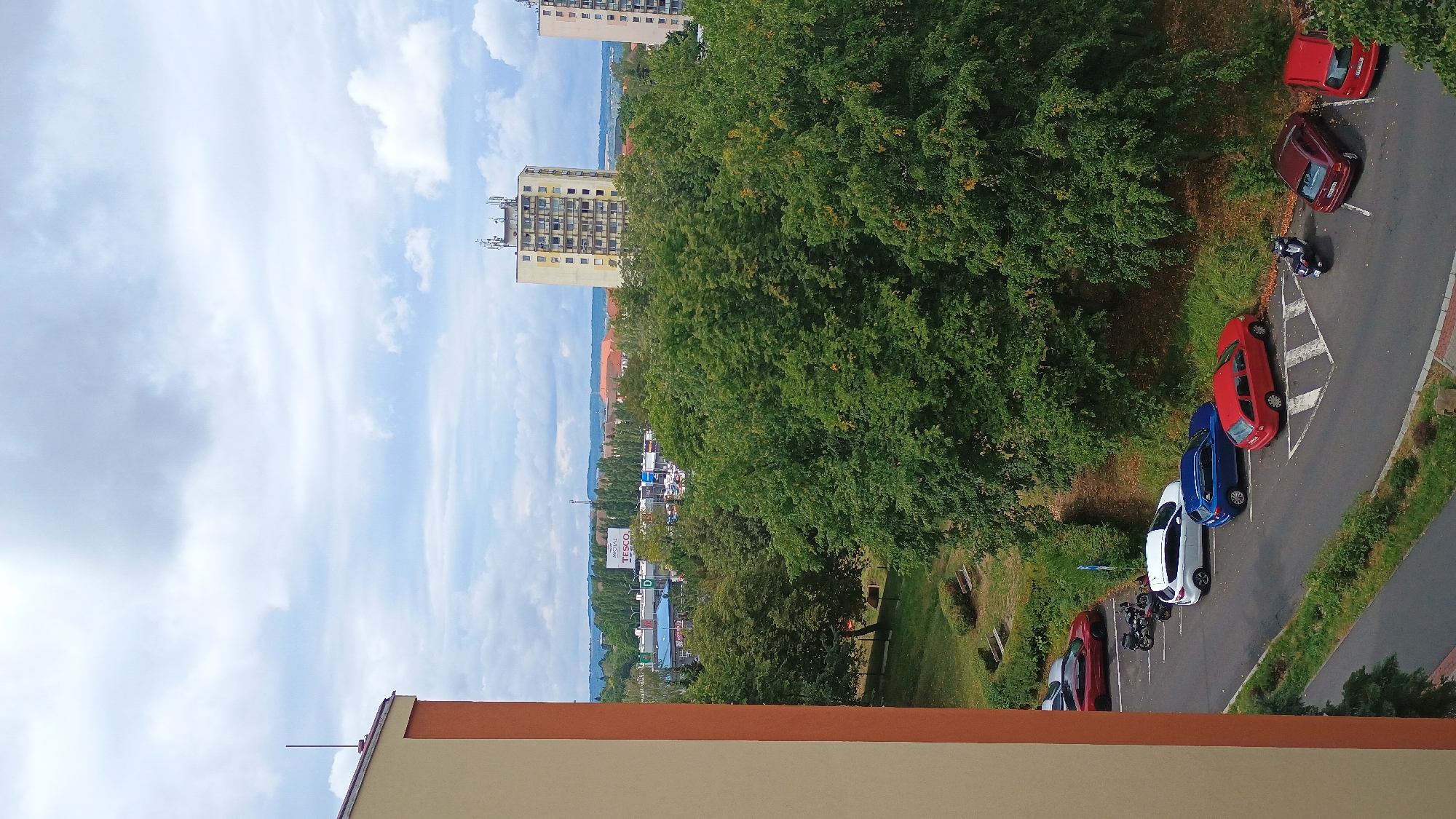The image captures a view from a building in Sokolov, Czechia. In the foreground, the side of a building with cream-colored walls and an orange trim is visible. Below, a parking lot contains several vehicles, including a red sedan, a white SUV, a blue sedan, a red hatchback, a maroon sedan, and a red sedan. A black motorcycle is also parked. Beyond the parking lot is a dense area of green trees. In the background, there are residential buildings, including a tall, light-yellow apartment building with numerous balconies and a smaller building to its right. Further in the distance, there is a commercial area with a sign that reads "TESCO." The sky is overcast with scattered clouds.
