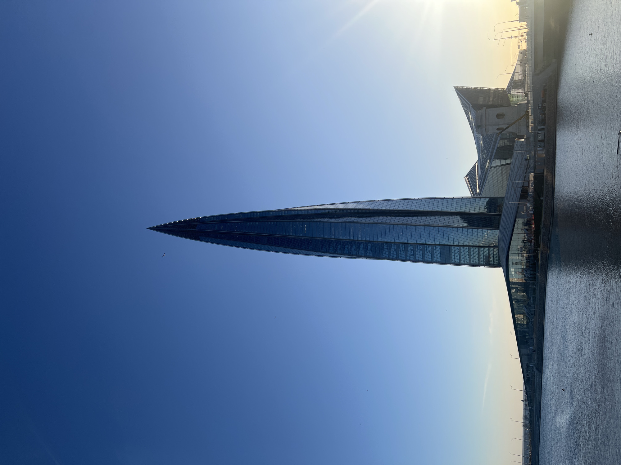 The image shows the Lakhta Center skyscraper in Saint Petersburg, Russia. It is a tall, modern glass building with a pointed apex, dominating the view against a clear blue sky. The sun is shining brightly, causing a glare at the top right of the image. To the right of the skyscraper, there are other structures and buildings along the waterfront, with a body of water visible on the right side. The water reflects the sky and the surrounding buildings. There are birds flying in the sky. The overall environment suggests a sunny day in a modern urban setting.