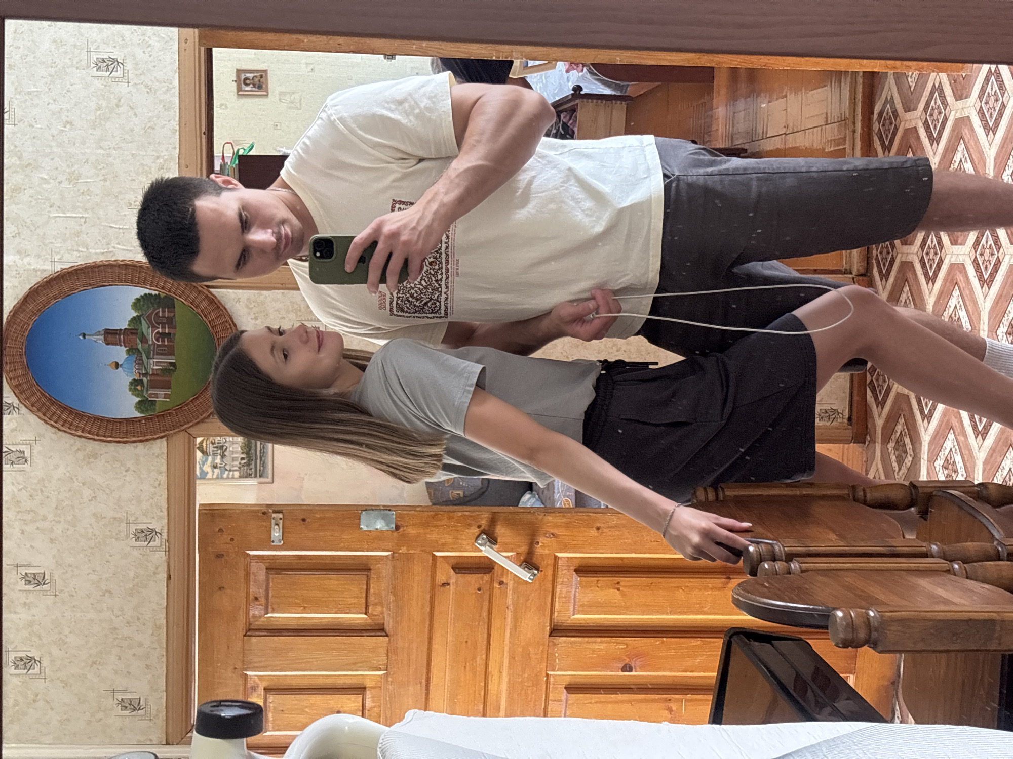 A man and a woman are taking a mirror selfie in an interior setting that appears to be a home in Krasnogorskoye, Russia. The woman is on the left, wearing a grey t-shirt and a black skirt. She is looking at the man and has her left hand resting on the back of a wooden chair. The man is on the right, wearing a white t-shirt and dark shorts. He is holding a smartphone in his right hand, taking the picture, and looking down at the phone. A white cord is hanging from the phone.
The background shows a wooden door with panels and a metal handle on the left. The wall above the door has patterned wallpaper and a framed oval painting of a church. To the right of the painting is a mirror reflecting the room. The floor is tiled with a geometric pattern. The overall environment suggests a modestly furnished living space.