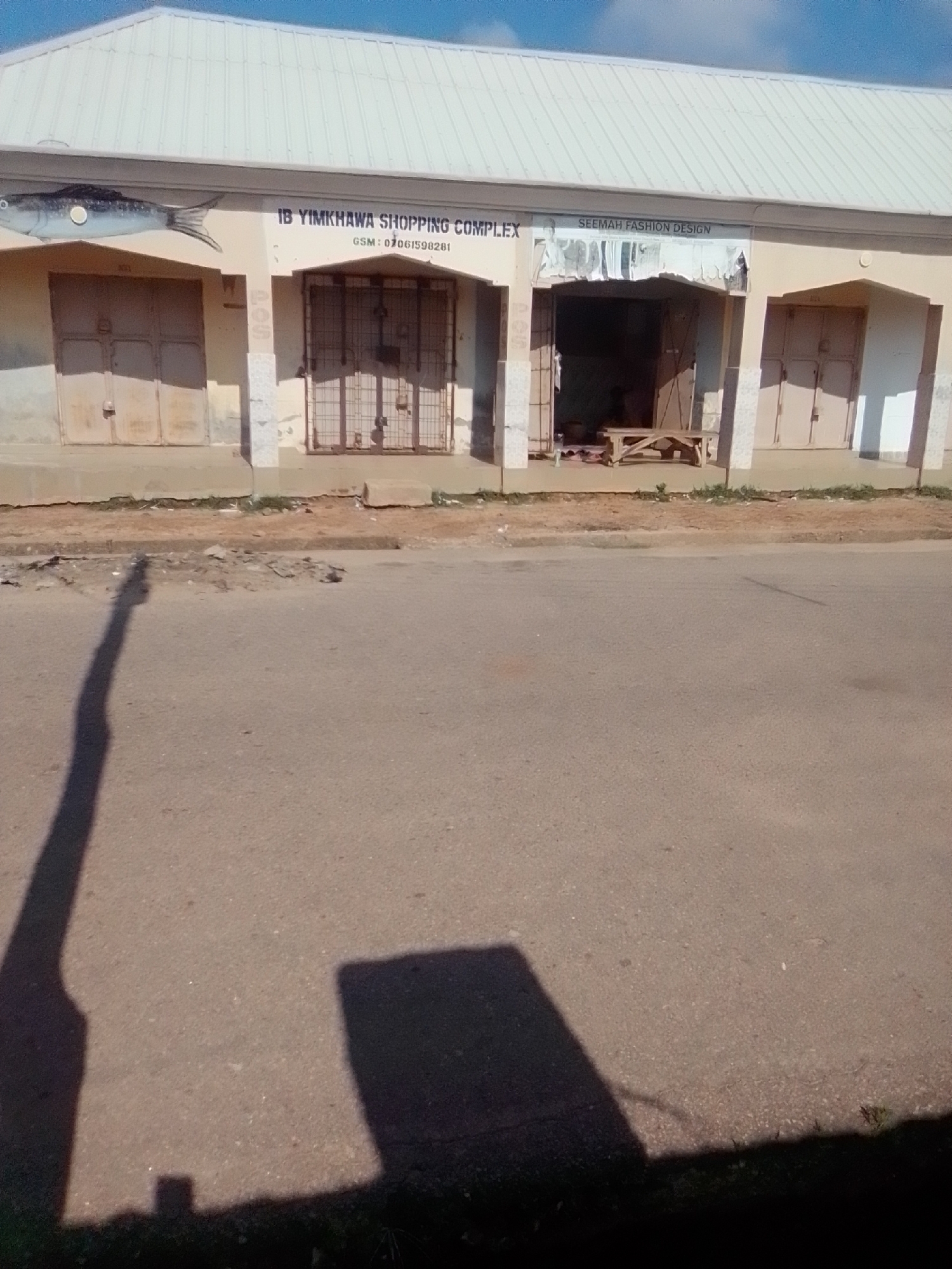 The image shows a low-angle view of a storefront in the city of Gombe, Nigeria, under a bright, cloudless sky. The storefront is part of a building with a corrugated metal roof. The facade is painted a light color, with darker trim around the windows and doors. A large, painted image of a fish is on the left side of the building. The storefront itself is divided into several sections, with large, metal-shuttered windows or doors. The central section of the storefront is open, revealing a simple wooden bench and some other objects inside. Above the central opening, a sign reads "SEEMAH FASHION DESIGN". Another sign to the left reads "IB YIMKHAWA SHOPPING COMPLEX" and includes a phone number. The ground in front of the storefront is paved, and there is a paved road in the foreground. Shadows of a person and an object are cast across the road, indicating the presence of the photographer and potentially other elements outside the frame. The overall impression is one of a commercial area, likely on a sunny day.