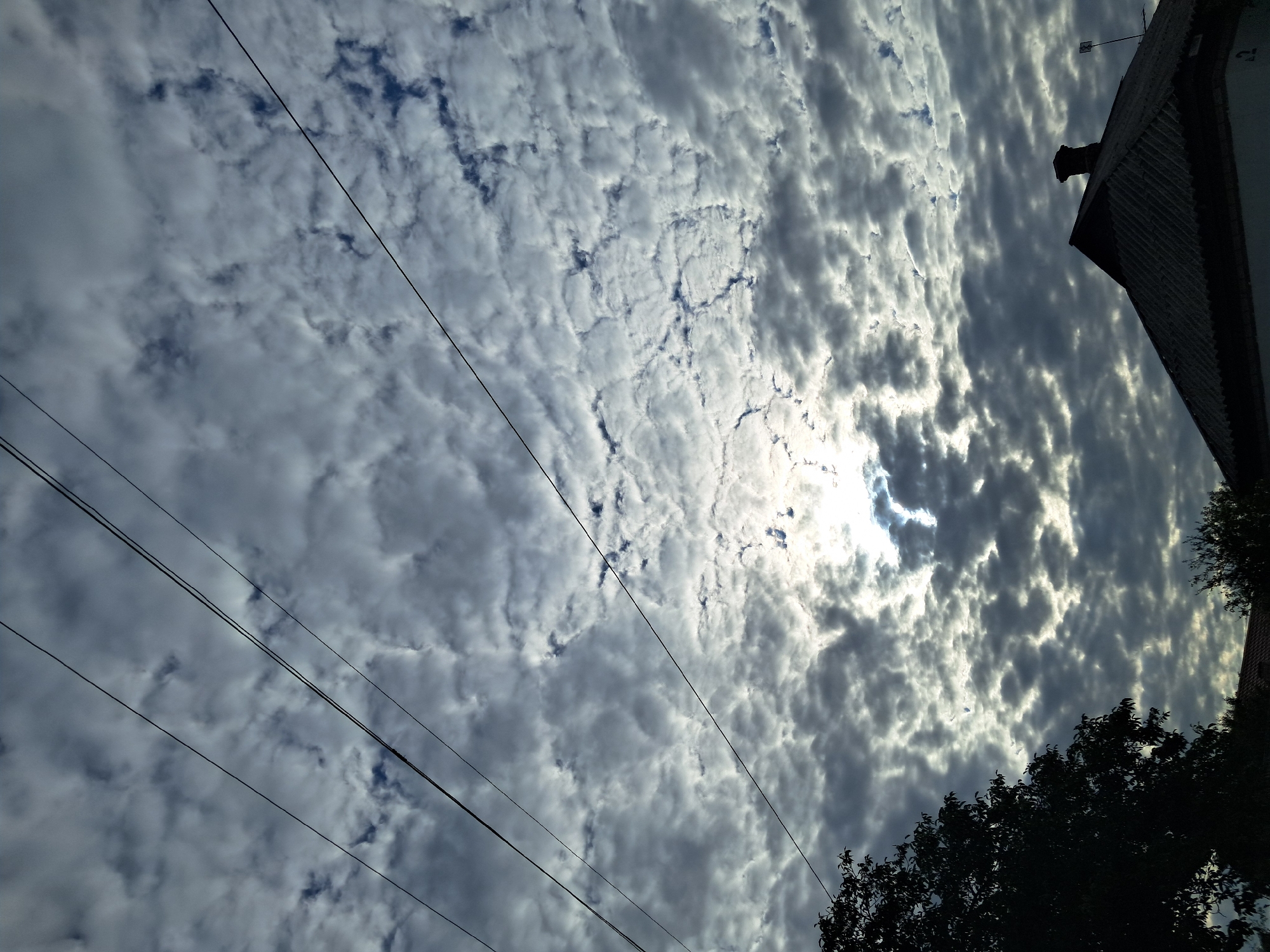 This is a vertical, medium-distance photograph taken from a low angle, looking up at a sky filled with a dramatic, textured cloud cover. The sun is partially obscured by the clouds, creating a bright, diffused light source that highlights the edges of the cloud formations. Several thin, dark power lines stretch diagonally across the upper portion of the frame. In the lower portion of the image, the silhouette of a dark, leafy tree is visible on the left, and the roof of a building, possibly a house, with a dark, corrugated surface and a chimney, occupies the lower right corner. The overall impression is of a cloudy, possibly overcast day with shafts of sunlight breaking through. The scene appears to be an outdoor urban or suburban environment, likely residential given the presence of the house and power lines. No people are visible in the frame. The lighting suggests it could be early morning or late afternoon.