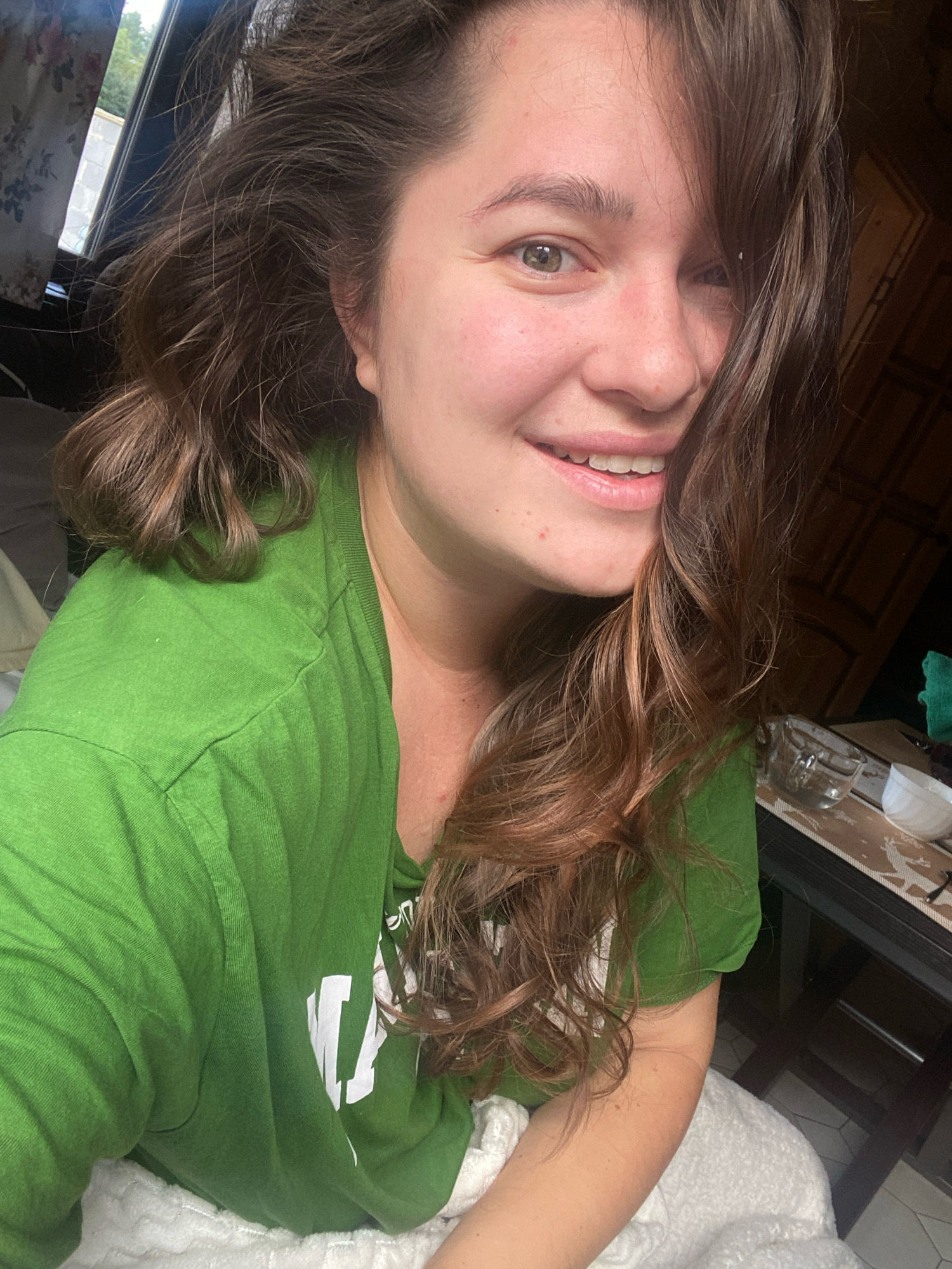 A young woman with brown wavy hair is taking a selfie indoors. She is wearing a green t-shirt and smiling. Her left eye is visible, and her right eye is partially obscured by her hair. The background shows a window with curtains, a dark wooden door, and a table with some objects on it, including a glass and a bowl. The image appears to have been taken in a residential setting. There are no visible indications of the city of Perm, Russia.