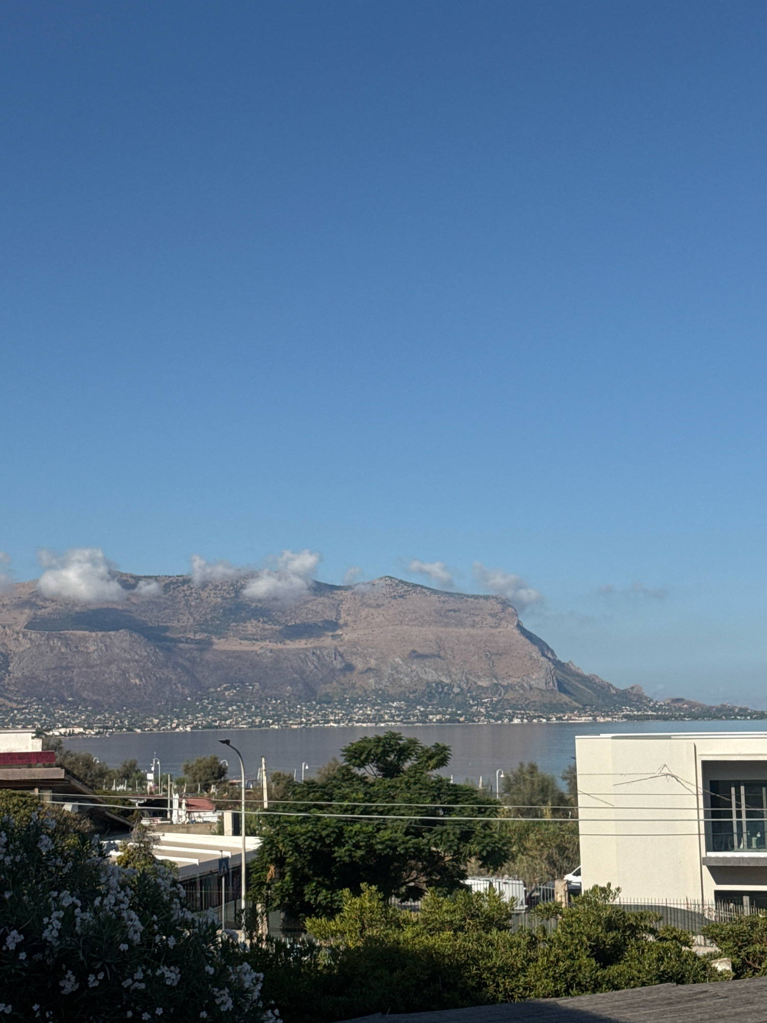 The image shows a scenic view of the Gulf of Castellammare from Capaci, Italy. In the foreground, there is a white building with a modern design and large windows. Behind it, lush green trees and bushes are visible, with white flowers in the lower left corner. Further in the background, the calm waters of the gulf stretch out, leading to a mountainous coastline dotted with houses. The mountains themselves are dry and rocky, with some scattered clouds clinging to their peaks. The sky is a clear, bright blue, indicating a sunny day. The scene conveys a sense of peaceful coastal living with a backdrop of natural beauty.