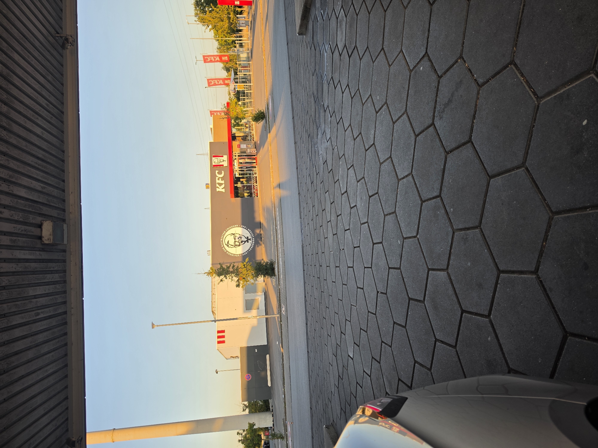 In Bischofsheim, Germany, a KFC restaurant is visible under a clear sky during what appears to be either sunrise or sunset, casting long shadows. The building features the iconic KFC logo and signage, with red and white accents. Red banners with the KFC logo are also displayed. In the foreground, a section of a silver car is visible, its reflection captured on its metallic surface. The ground is paved with dark gray hexagonal tiles. A tall white pillar is present on the left side of the image. The scene suggests an outdoor dining or drive-thru area associated with the fast-food establishment.