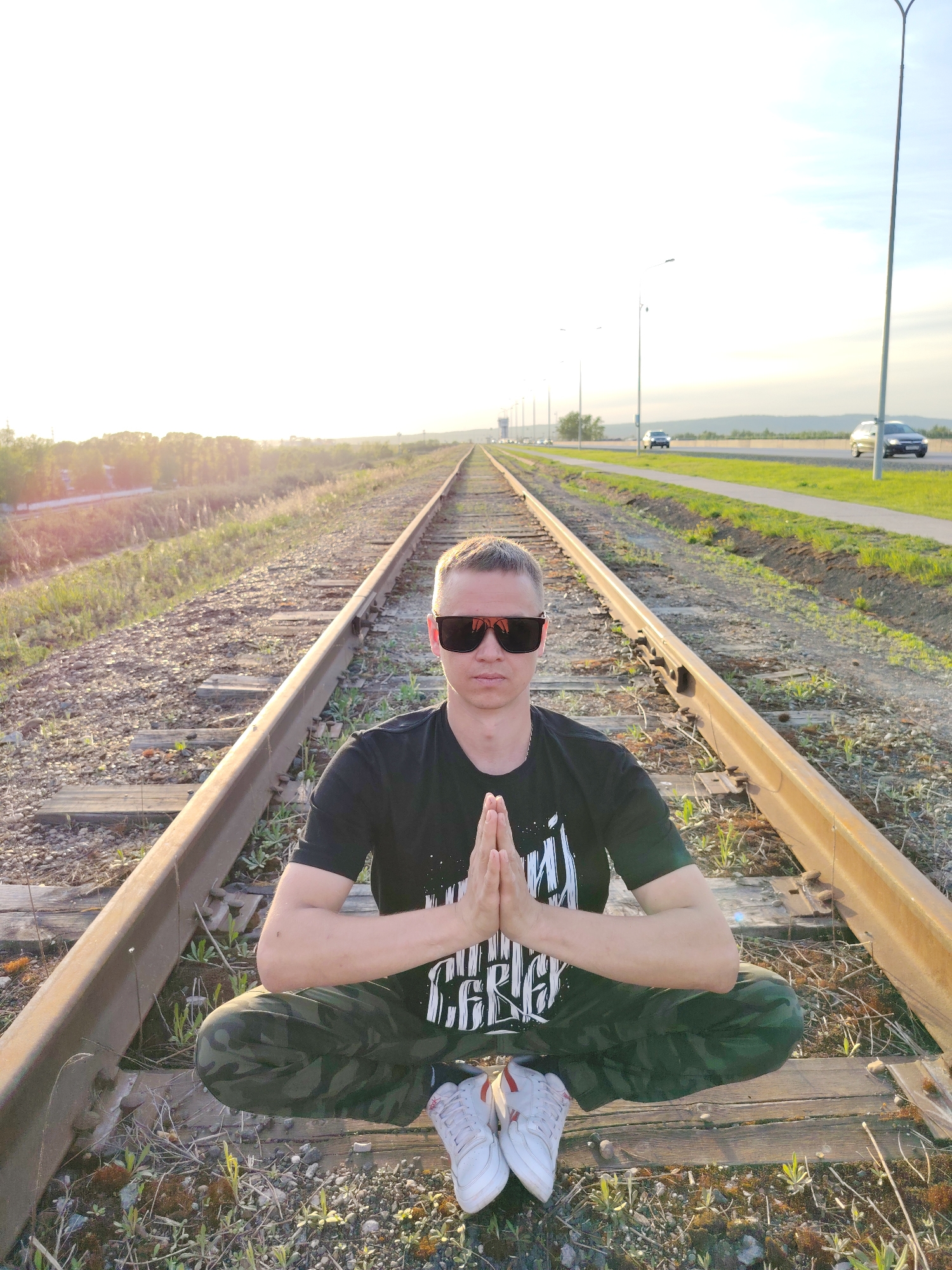 A medium shot depicts a fair-skinned man with short blond hair sitting cross-legged on a railroad track, hands pressed together in a gesture of prayer or meditation. He wears sunglasses, a black t-shirt with a white graphic design, and camouflage pants, with white sneakers visible at the bottom of the frame.
The location appears to be in Chaikovsky, Russia, suggested by the industrial and transportation landscape.
The railroad tracks extend into the distance, creating a vanishing point perspective. The environment includes roadside vegetation, a multi-lane highway with cars, and lampposts, indicating a modern urban area. The sky is bright, suggesting it might be early morning or late afternoon. The overall atmosphere is serene, contrasting the calmness of the man with the busy transportation infrastructure.