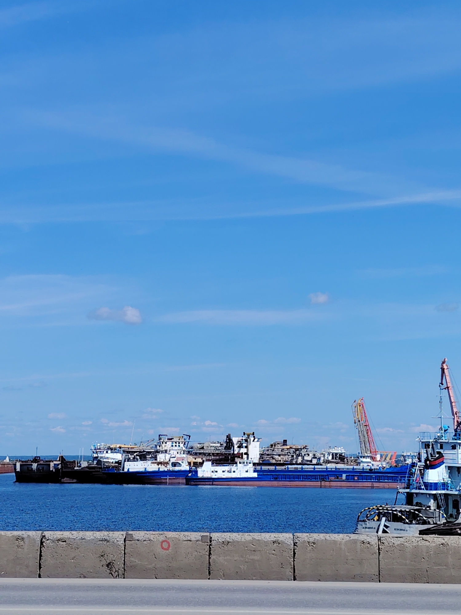 The image depicts a section of the Kama River port in Chaikovsky, Russia, on a clear, sunny day. The foreground features a concrete barrier, possibly a seawall or dock edge, with a road or walkway visible in front of it. Beyond the barrier, the blue waters of the Kama River are calm.
In the mid-ground, several barges and ships are moored. One prominent, long blue barge is laden with what appears to be industrial equipment or scrap metal. Several other vessels, a mix of white and blue, are also docked, some appearing to be smaller tugboats or support craft. In the background, a large red and yellow construction crane is visible, along with other industrial structures. The sky is a bright, clear blue with a few wispy white clouds. No people are visible in the scene. The overall impression is one of a working port under a pleasant sky.