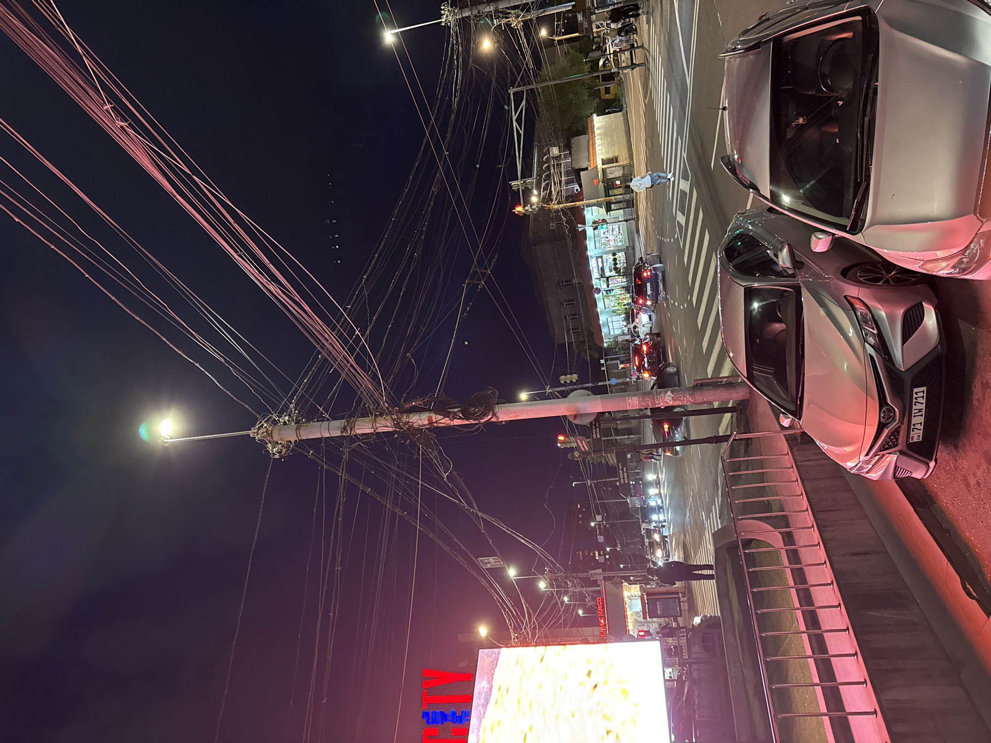 The image shows a nighttime scene in Yerevan, Armenia. A tall utility pole is heavily laden with tangled wires. A street light extends from the top of the pole. Below the pole, there is a bright billboard with the word "FLY" in large red letters, and some other text.
To the right of the image, two parked silver cars are visible. One car's license plate is partially visible and reads "71 IW 711." A concrete wall with a metal railing runs alongside the cars. Across the street, traffic is visible with red and white lights, along with several buildings and streetlights. A person is standing across the street. The sky is dark.