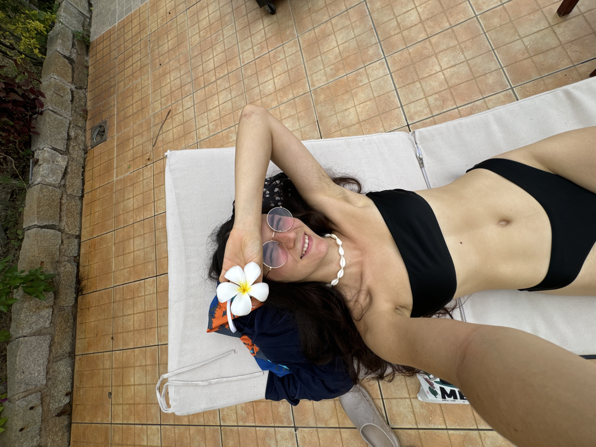 An overhead shot captures a woman lying on a lounge chair in an outdoor setting, taking a selfie. She is smiling broadly, conveying a sense of happiness and relaxation. Her long, dark hair is spread out around her head, and she is wearing a black bikini top and bottoms, along with a necklace made of white cowrie shells and round, reflective glasses.
Her left arm is bent, holding a white plumeria flower near her temple, while her right arm extends towards the viewer, likely holding the camera. Beneath her head, a dark blue garment with an orange pattern is visible. Near her feet, a light-colored slipper or sandal rests on the tiled floor, next to an item that displays the letter "M" with further blurred text.
The ground around the lounge chair is covered with small, square, brownish-orange tiles. To the left, a rustic stone wall or pillar is visible, with green and reddish foliage peeking out from behind it. A circular drain cover is also present on the tiled surface. The scene suggests a warm, sunny day in a tropical or subtropical location, fitting the given context of Phu Thuy Ward, Vietnam.