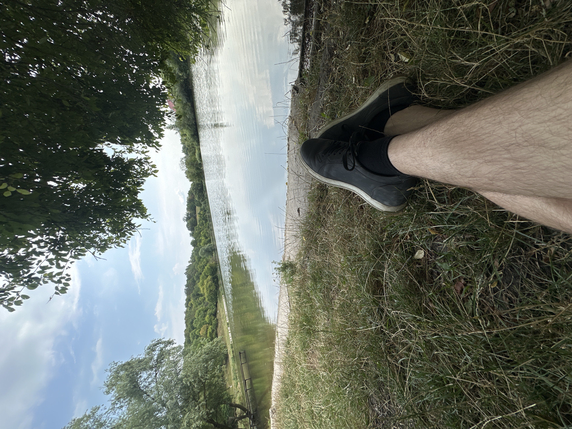 A person's legs are crossed and resting on the grass in the foreground, with black sneakers and dark socks visible. Beyond the grass, a calm body of water reflects the sky, which is partly cloudy and blue. Lush green trees line the opposite bank of the water, with a gentle slope leading up to a treeline. A wooden structure, possibly a pier, extends into the water from the left bank. The overall scene depicts a peaceful outdoor setting by a lake. The location is identified as Makiv, Ukraine.
