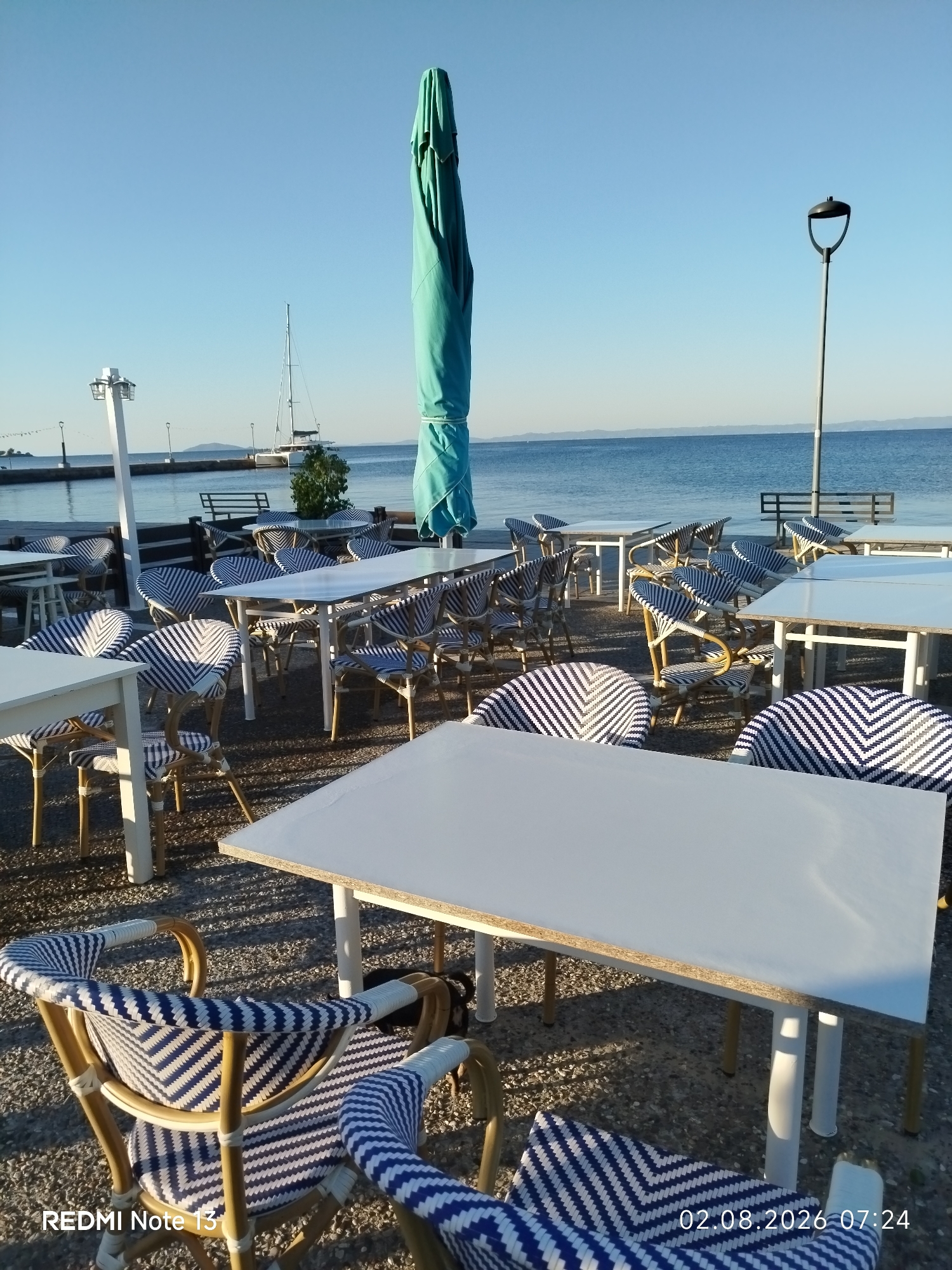 The image depicts an outdoor seating area of a cafe or restaurant situated on a waterfront, likely in the Sithonia Municipal Unit, Greece, given the visible timestamp. The foreground is occupied by several white-topped tables and chairs with a distinctive woven pattern of blue and white stripes on their seats and backs. These chairs appear to be made of rattan or a similar material. In the background, the turquoise water of the sea stretches to the horizon under a clear blue sky.
A tall, folded turquoise umbrella stands prominently near the center-right of the frame. Beyond the seating area, a stone pier or walkway is visible, with a few street lamps and a modern-looking white pole with lights. A white sailboat with tall masts is anchored or docked in the water behind the pier. The ground is covered with small grey pebbles. The overall scene suggests a peaceful morning or early evening setting, with the sun casting distinct shadows from the tables and chairs onto the ground. No people are visible in the frame. The timestamp "02.08.2026 07:24" is overlaid in the bottom right corner, and "REDMI Note 13" is in the bottom left corner.