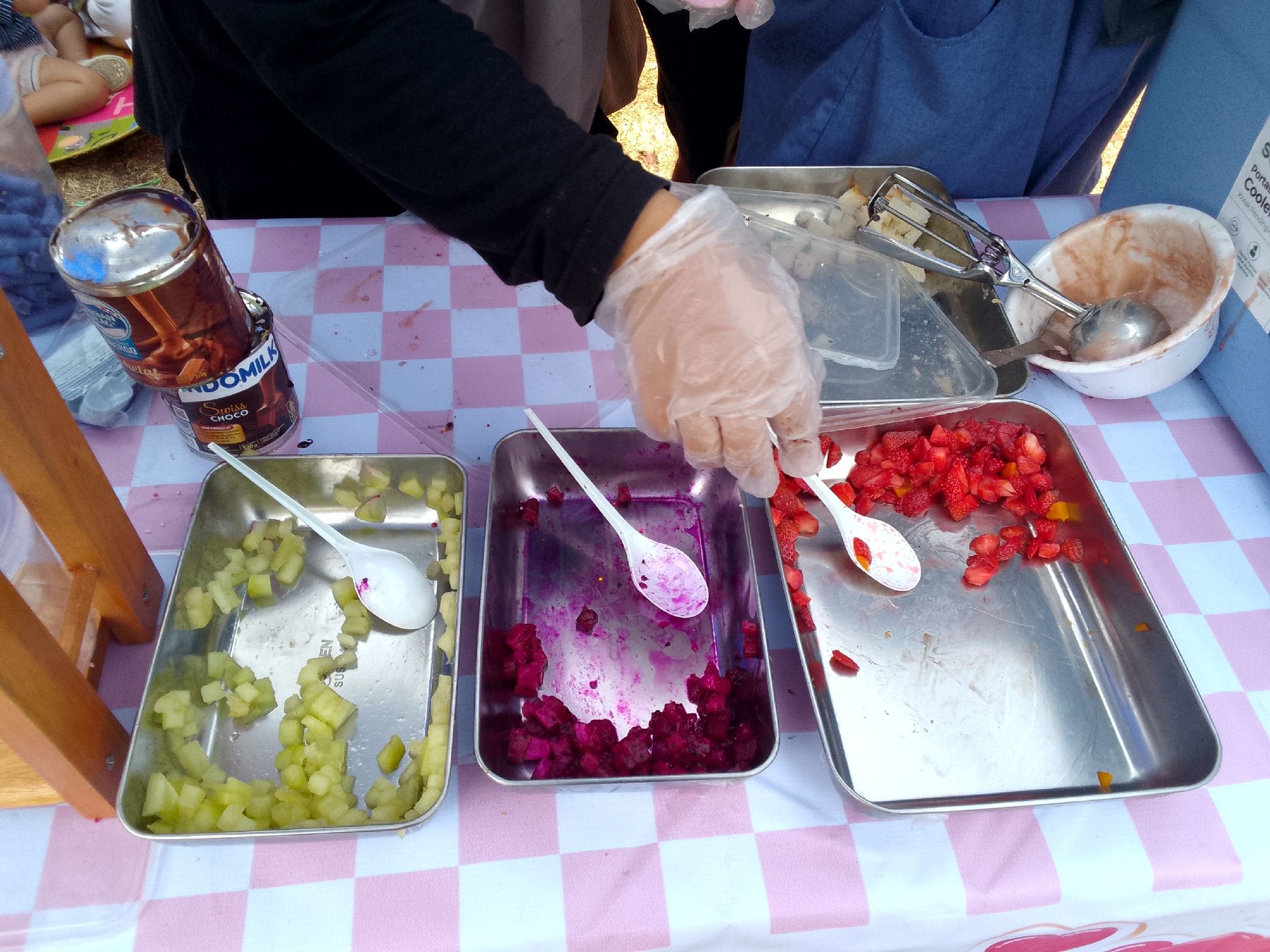The image depicts an outdoor food preparation scene, likely at a public event or market stall in Mandalawangi, Indonesia. In the foreground, three metal trays are arranged on a pink and white checkered tablecloth. The leftmost tray contains diced green fruit, possibly apples or pears, with a white spoon resting inside. The middle tray holds chopped dark red fruit, possibly dragon fruit, with purple residue staining the spoon within. The rightmost tray is filled with chopped strawberries, and a white spoon with red fruit residue is visible. A person's hands, wearing clear plastic gloves, are visible reaching towards the middle tray, suggesting they are in the process of preparing or serving the fruit.
Behind the trays, a metal can with a chocolate milk advertisement is partially visible, standing next to a blue cooler. An ice cream scoop rests on a clear plastic container. In the background, the legs of several individuals are visible, indicating the presence of customers or other event participants. The ground appears to be dirt or grass, further suggesting an outdoor setting. The overall impression is one of a community event or casual food service operation in Mandalawangi, Indonesia.