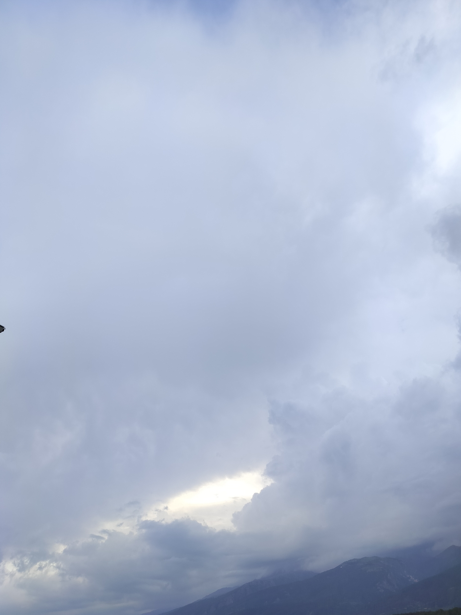 The image presents a landscape view dominated by a vast, cloudy sky over mountains, likely in Gorno Sahrane, Bulgaria. The upper two-thirds of the frame are filled with dense, varied cloud formations ranging from light blue-grey to darker, heavier shades of grey. A prominent feature is a bright, luminous patch of white-yellow light breaking through the clouds in the lower-central and right portions of the sky, indicating the sun's position or a significant clearing. This creates a strong contrast between the radiant light and the surrounding darker cloud masses.
In the bottom third of the image, the silhouettes of mountains are visible, their dark forms partially obscured by lower-lying clouds and atmospheric haze, suggesting a grand and rugged topography. The overall weather condition appears overcast but dynamic, with the sun attempting to pierce through, possibly indicating late afternoon or early morning. A tiny, dark, indistinct object or structure is partially visible at the far left edge of the frame, but it is largely out of focus and unidentifiable. No text is visible in the image.