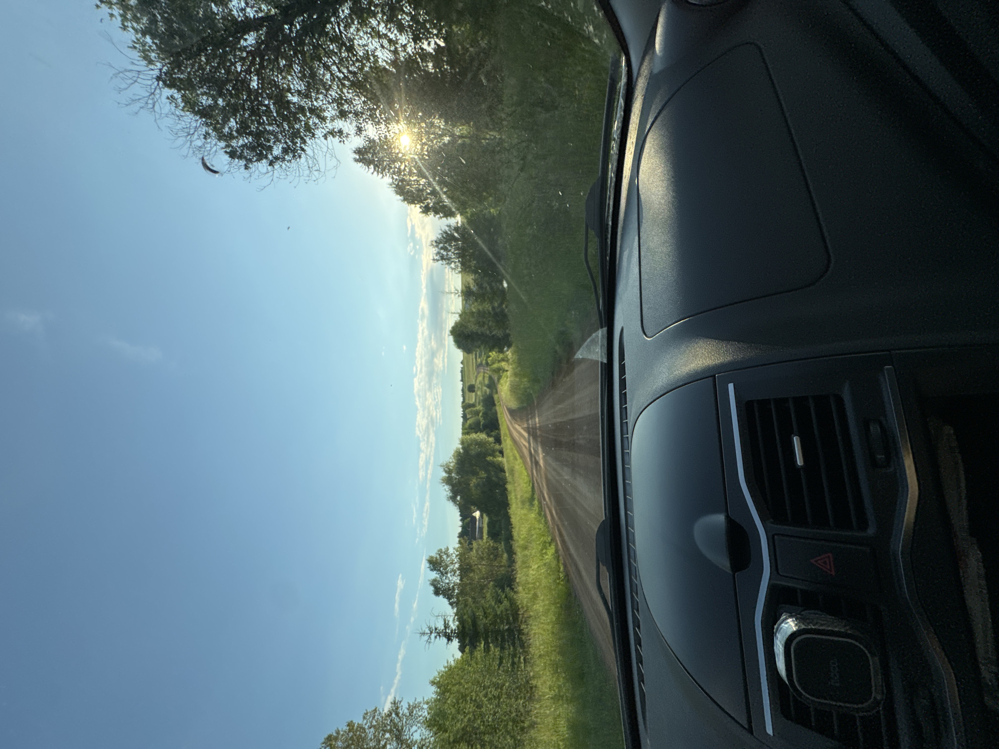 This is a first-person view from inside a car driving down a dirt road in Amaneevo, Russia. The sun is low in the sky, casting a warm glow and creating lens flare through the trees on the right side of the image. The road is flanked by lush green grass and trees. In the distance, there are hints of houses, suggesting a rural setting. The car's dashboard is visible in the foreground, with a phone mount attached to the left side. The sky is clear blue with a few wispy clouds.