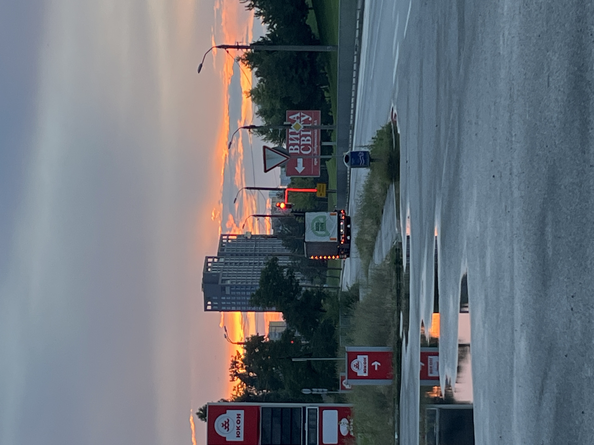 A wide shot captures an urban scene at sunset in Dnipro, Ukraine. The sky is filled with vibrant orange and pink clouds, indicative of the setting sun. In the foreground, a wet, gray asphalt surface reflects the colorful sky and a red sign with a white arrow and the Cyrillic word "ЮКОН" (Yukon). To the left, a tall red sign with the "ЮКОН" logo stands next to a gas pump display. In the middle ground, a semi-truck with a white trailer, bearing a green logo with Cyrillic text, is driving on a paved road. A red traffic light is visible to the left of the truck. Further back, a modern, tall building with a dark facade and numerous windows dominates the skyline, with other buildings and trees scattered throughout the urban landscape. To the right of the truck, a prominent red sign with white Cyrillic text that reads "ВИЛА СВІТУ" (Vila Svitou), meaning "Village of Light," is positioned beside a yield sign. A street lamp is visible near the sign. The environment appears to be a roadside area with some grassy patches and a concrete barrier along the road.