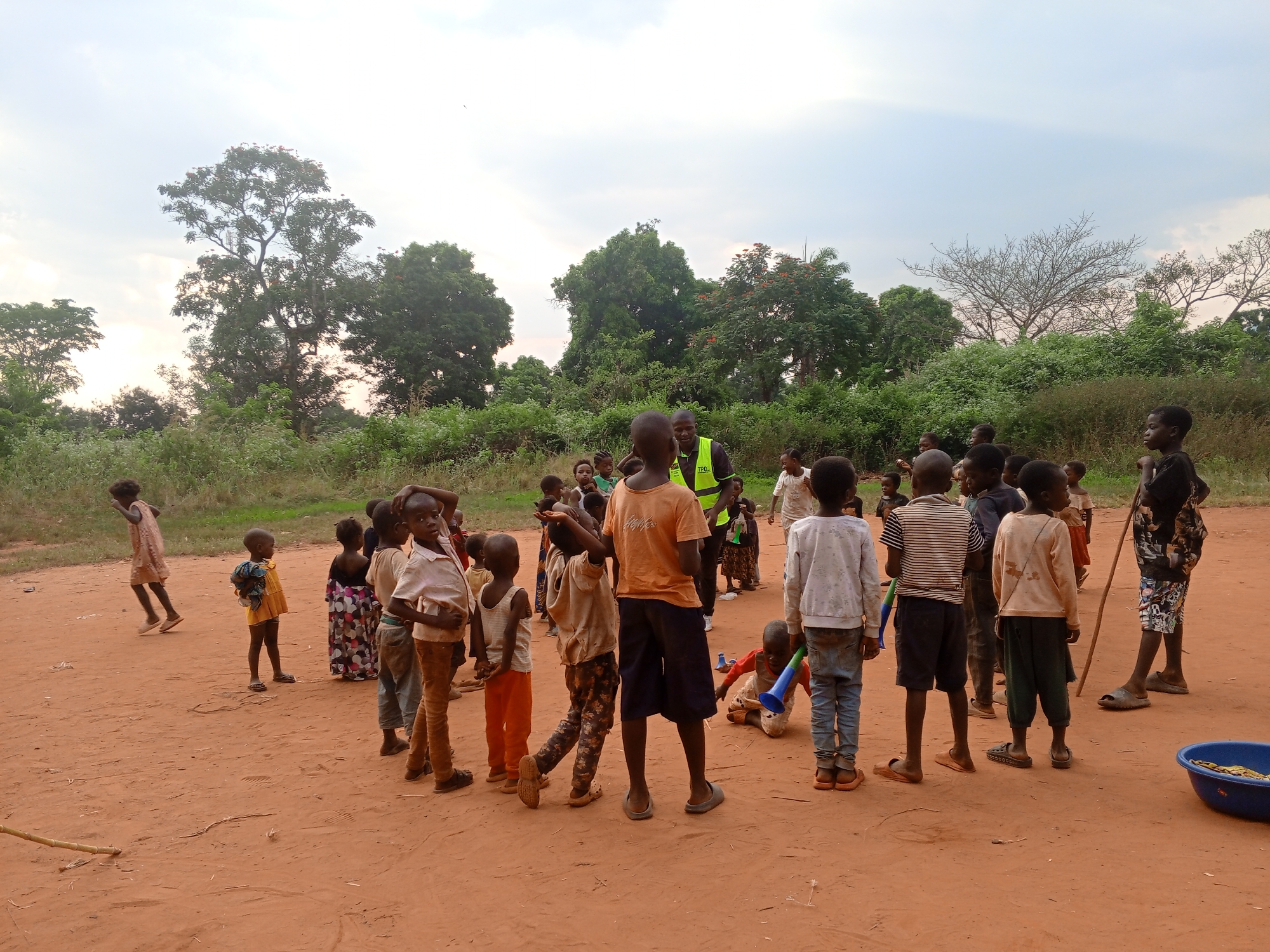 A wide-angle shot depicts a group of children and an adult in Ngalula, Democratic Republic of the Congo. The setting is an open, dirt area with trees and vegetation in the background under a cloudy sky. Approximately twenty children are present, wearing various casual clothes, some barefoot, others in sandals. They stand and interact, with one running towards the group, and another kneeling with a blue plastic horn. An adult wears an orange t-shirt and a neon yellow vest, standing in the center of the children. A blue basin sits on the ground towards the right edge of the frame.