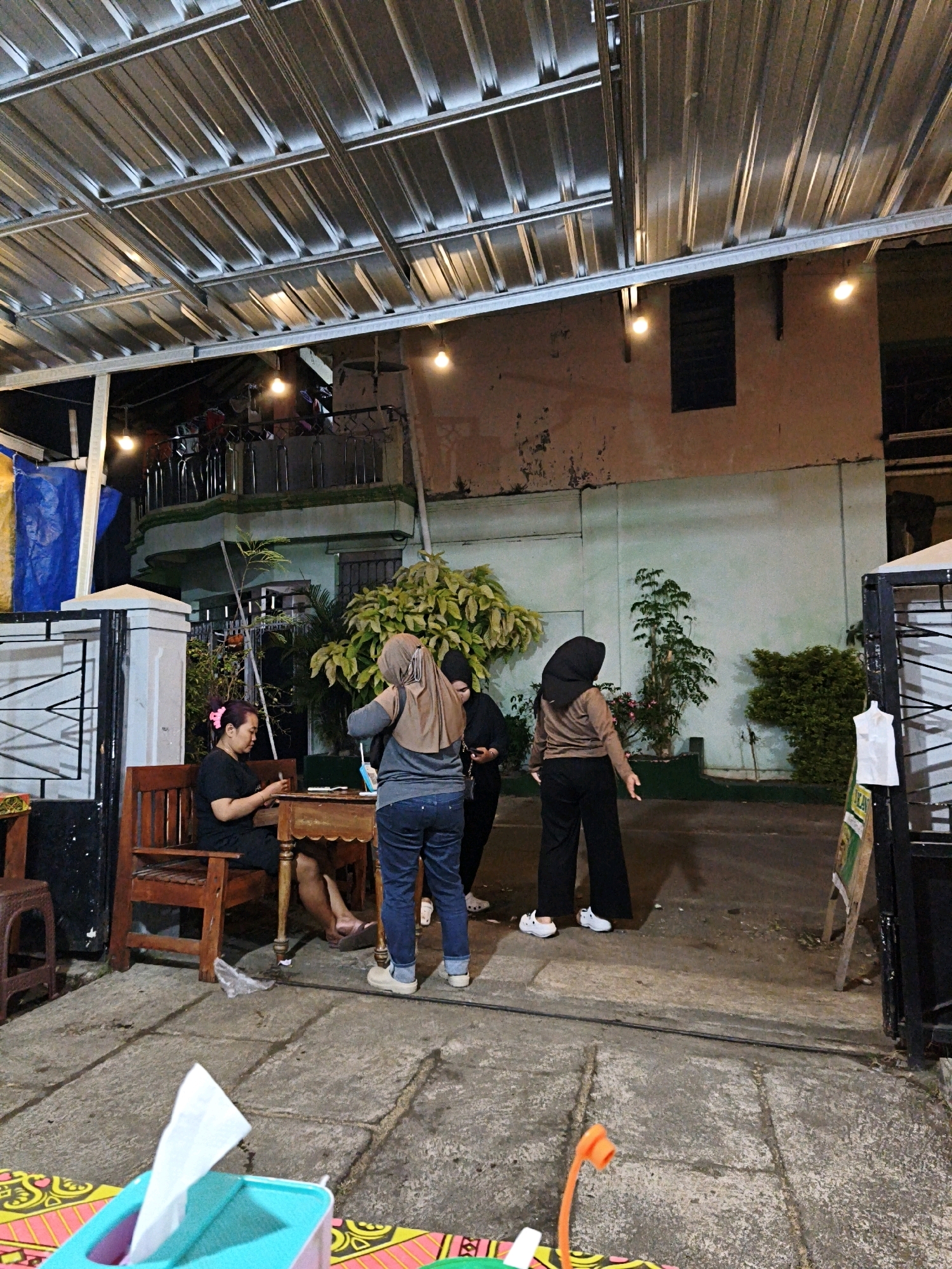The image captures a scene at night in front of a building in the city of Surakarta, Indonesia. A corrugated metal roof with exposed beams and incandescent lights covers an outdoor seating area. A woman wearing a black t-shirt and dark shorts with a pink hair tie sits on a wooden bench at a wooden table, looking down at her hands, which hold a small object, possibly a phone. Another woman, wearing a light brown hijab, grey t-shirt, and blue jeans, stands facing away from the camera, adjusting something in her hand. A third person, wearing a black hijab and black clothing, stands to the right of the second woman, partially obscured and also facing away. The building in the background has a light green facade with peeling paint and a dark rectangular window. Several plants, including a large leafy shrub and a smaller tree, are visible. The ground is paved with stone tiles and concrete. A black metal gate with white accents stands to the left of the frame. In the foreground, a portion of a colorful mat with a tissue dispenser is visible.