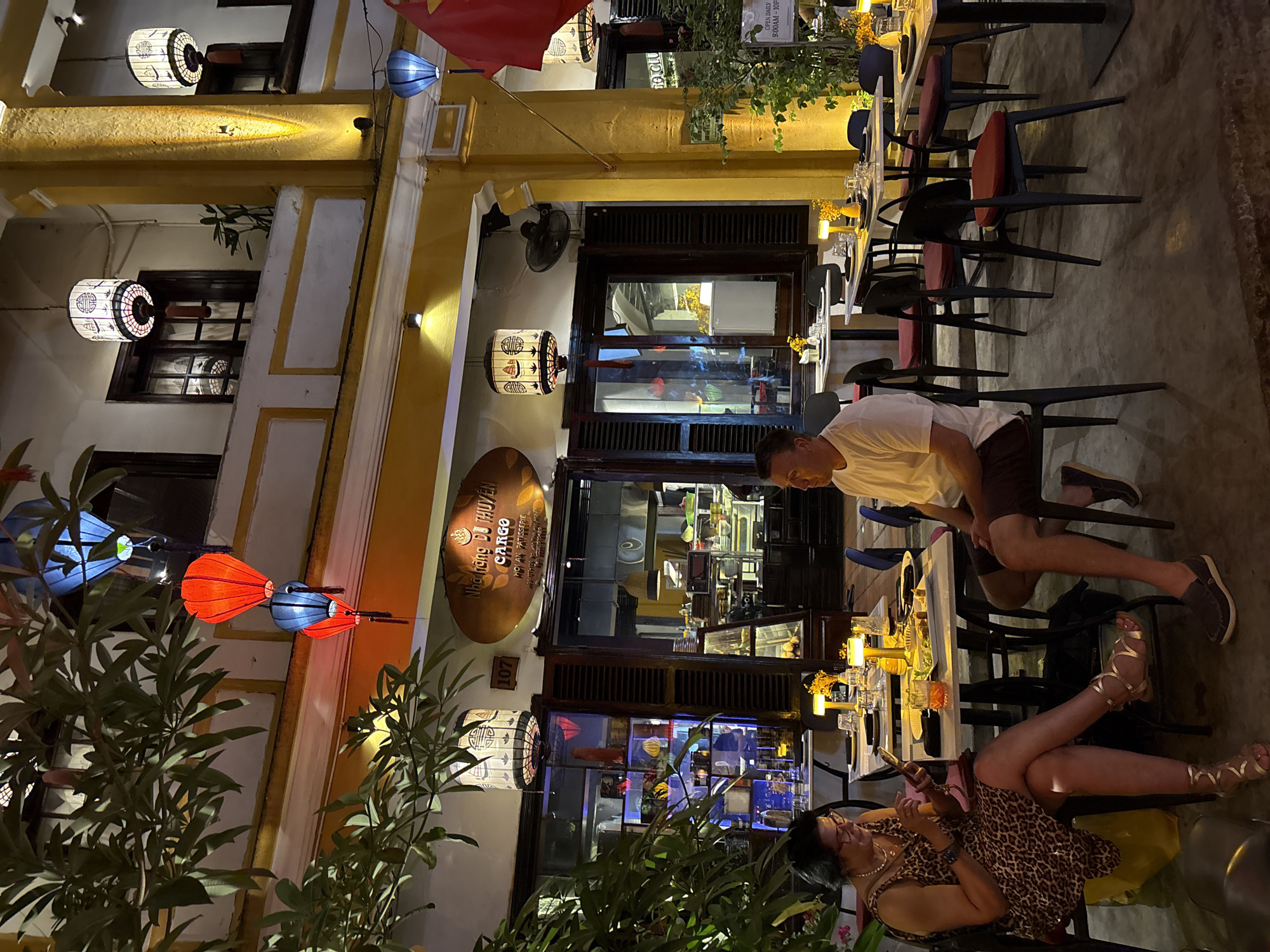 Here's a description of the image:
The image captures an outdoor dining scene in Hội An Ward, Vietnam at night. The foreground is partially obscured by foliage. Two people are seated at a long table set for dining. A woman wearing a leopard-print dress is in the foreground, looking at her phone. A man in a white t-shirt and dark shorts stands near her.
Behind them, the facade of a restaurant with golden yellow walls is visible. Lanterns in various colors (blue, red, white) decorate the exterior. The restaurant's signage reads "Nhà hàng DỠ THUYÊN CARGO HOI AN PATISSERIE," with the number "107." The restaurant has shuttered windows that reveal interior views, including a kitchen or bar area. Chairs are arranged along the table. A red flag waves above.