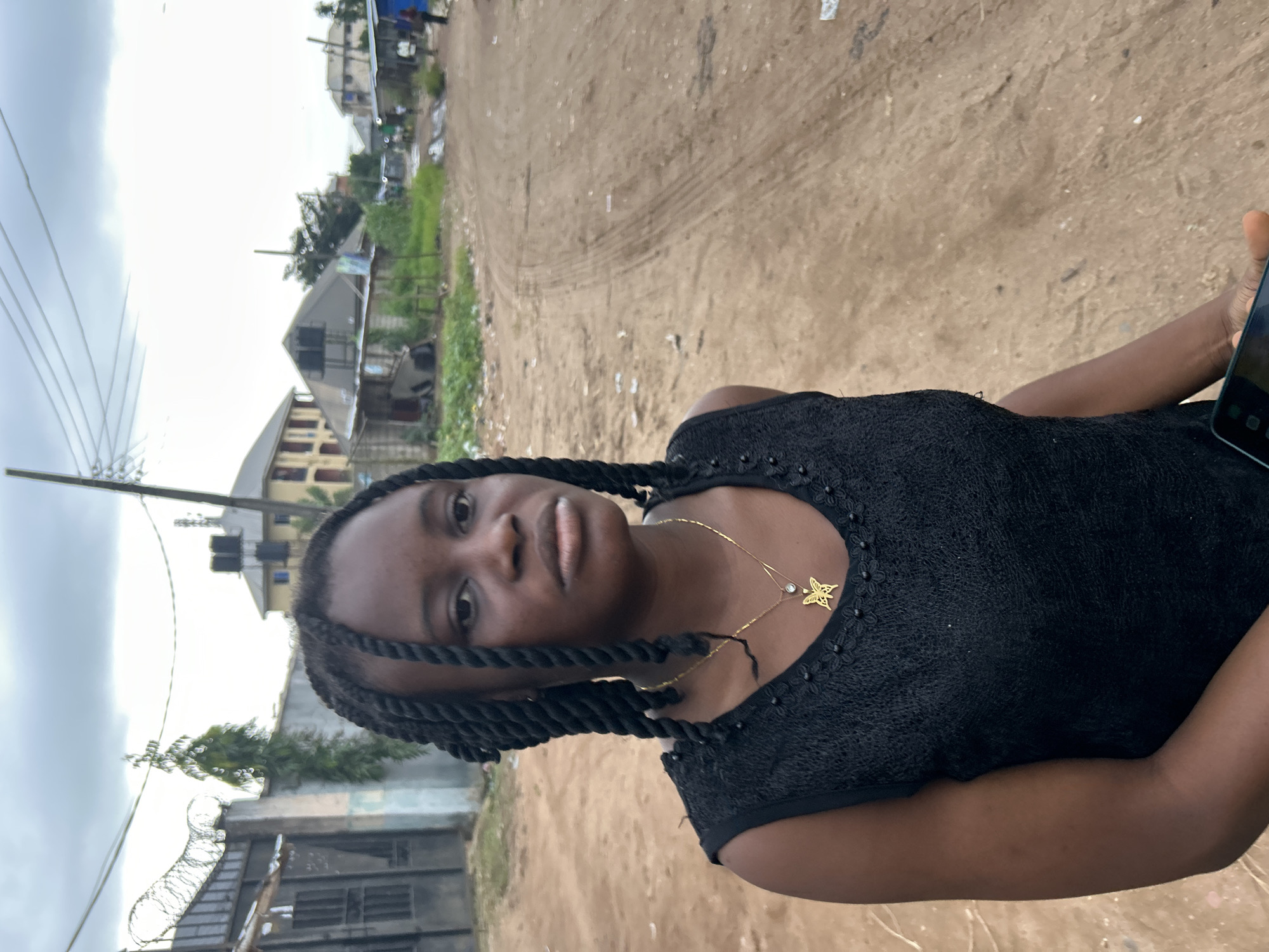 A young woman with dark, braided hair is the central subject of this vertical, medium shot image. She is wearing a black top adorned with small embellishments around the neckline. A delicate gold necklace with a butterfly pendant rests on her chest. Her gaze is directed slightly away from the camera, her expression neutral. Her right arm is visible, holding a dark-colored mobile phone.
The background suggests an outdoor setting in what appears to be a somewhat rural or developing area. A dirt road, marked with tire tracks, dominates the midground and background. Scattered debris is visible on the ground. To the left of the woman, buildings with visible windows and roofs, some appearing to be residential, are present. There are also power lines and poles. A section of barbed wire fencing is also visible in the lower left corner.
The lighting suggests it might be daytime, possibly overcast given the bright, washed-out sky. The overall impression is of a candid or casual moment captured outdoors. No visible text is present in the image. The location context is Ihiagwa, Nigeria.