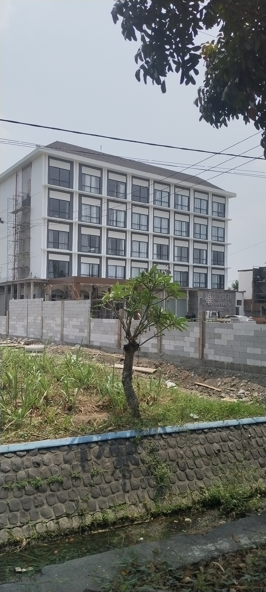 The image shows a modern, multi-story hotel building with a white facade and dark gray window frames. The building appears to be under construction or recently completed, with scaffolding visible on the left side. In front of the hotel, a low wall constructed from concrete blocks separates the property from a grassy area with a young tree. A water channel runs along the bottom of the image, bordered by a stone retaining wall. The scene is set against a bright, overcast sky. The hotel has signage that reads "LYNN HOTEL". The location is identified as Sengon, Indonesia.