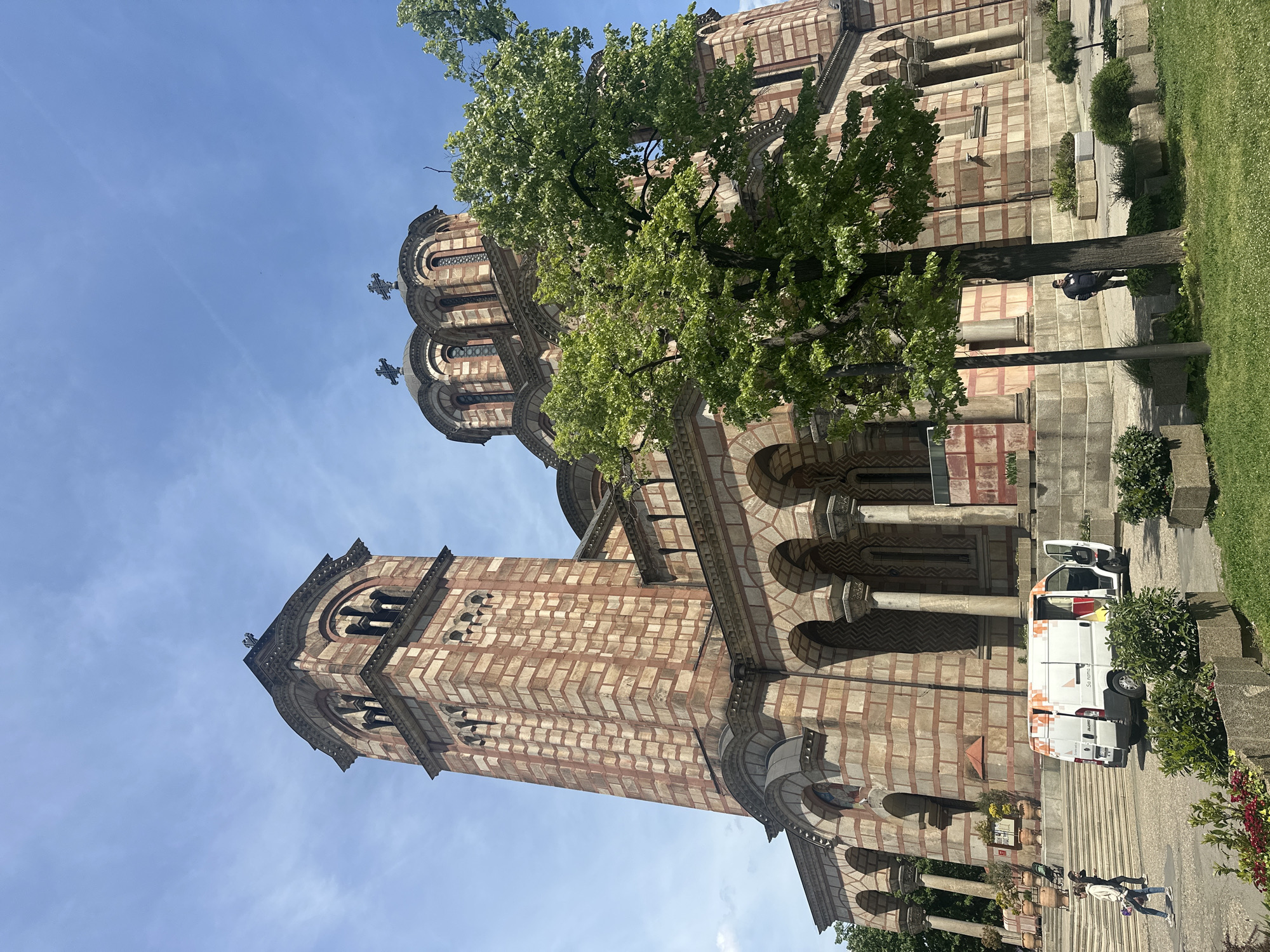 The image shows the Church of Saint Mark, a Serbian Orthodox church in Belgrade, Serbia, situated in the Palilula Urban Municipality. The church is a large structure with a prominent bell tower and multiple domes topped with crosses. The exterior is characterized by a pattern of alternating light and dark brown bricks. A tree partially obscures the view of the church. A white van with orange graphics is parked near the church, on a set of stairs. People can be seen walking on the sidewalk near the church. The sky is blue.