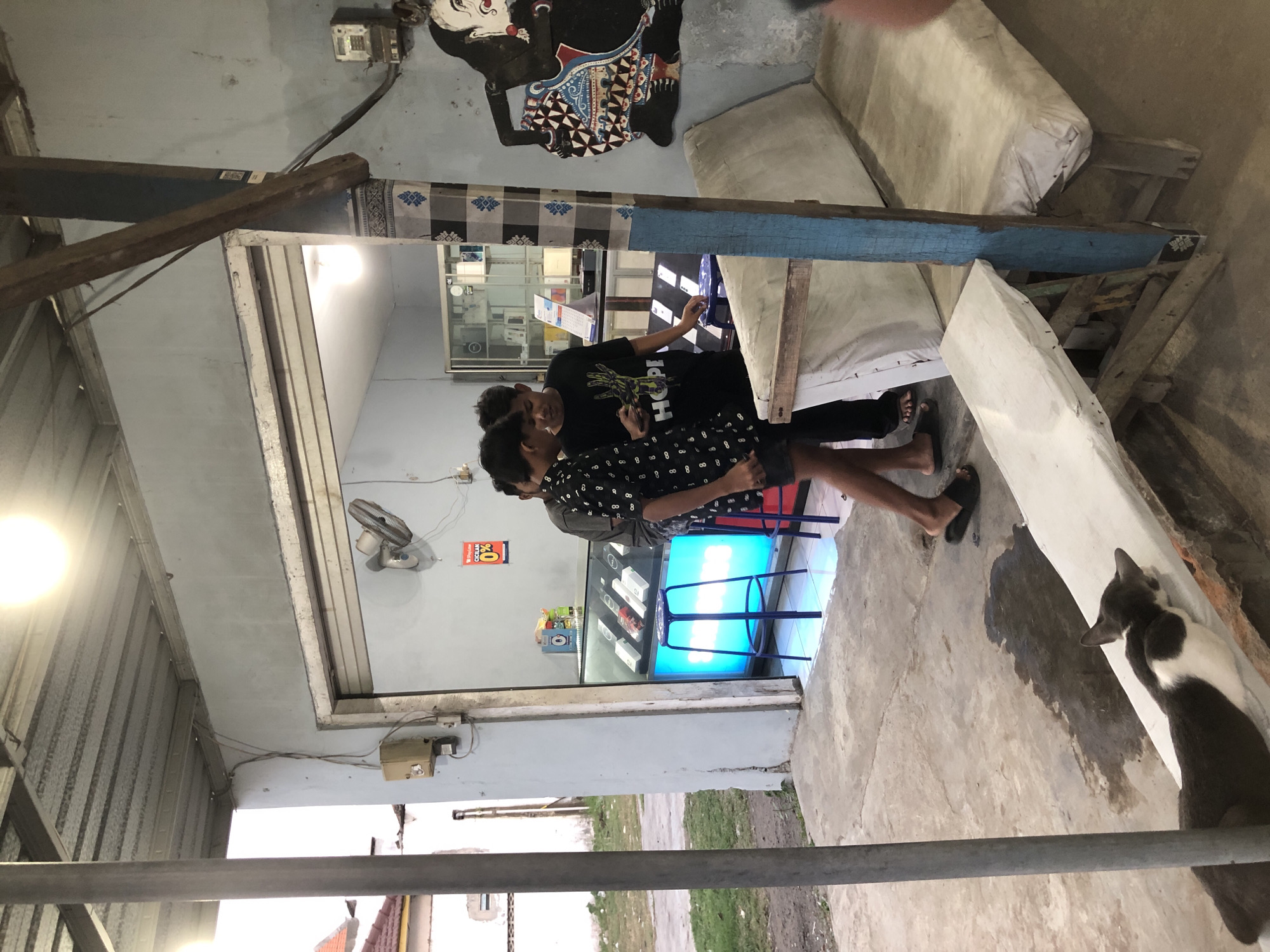In front of a small electronics store in Kesambi, Indonesia, two young males are engaged in conversation while looking at a mobile phone. The store is open to the street, with a display case inside filled with mobile phones. Two blue stools are positioned in front of the display. To the right of the store entrance, a large, traditional shadow puppet figure is painted on the wall. A tattered, padded bench sits outside the store. In the foreground, a cat lies on a low ledge. The overall environment appears to be a modest, open-air establishment in a somewhat unkempt urban setting, possibly a marketplace or a side street.