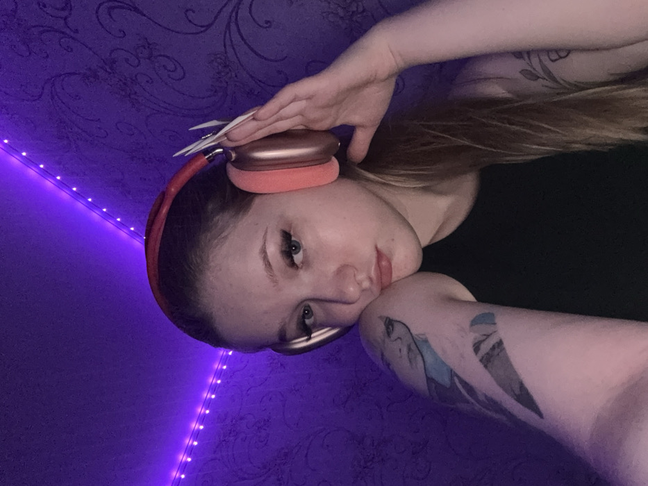 An indoor portrait depicts a young female subject. She possesses light skin and long, light brown hair. She is wearing a black sleeveless top and rose gold/pink over-ear headphones. Her left arm is bent, with her face resting against her left bicep. Visible on her left arm is a multi-colored tattoo depicting a stylized female face and geometric designs. Her right hand, adorned with long, sharply pointed white artificial nails, is positioned with fingers touching the right ear cup of the headphones. A tattoo resembling a leafy vine is partially visible on her right upper arm. The subject maintains direct eye contact with a subtle pout. The background consists of a wall covered in dark-toned, patterned wallpaper, likely with an embossed floral or damask design. The scene is illuminated by strong purple ambient light originating from a string of LED lights visible in the upper left corner of the frame. The specific location within the city of Saratov, Russia, cannot be ascertained from the visual evidence presented in the image.