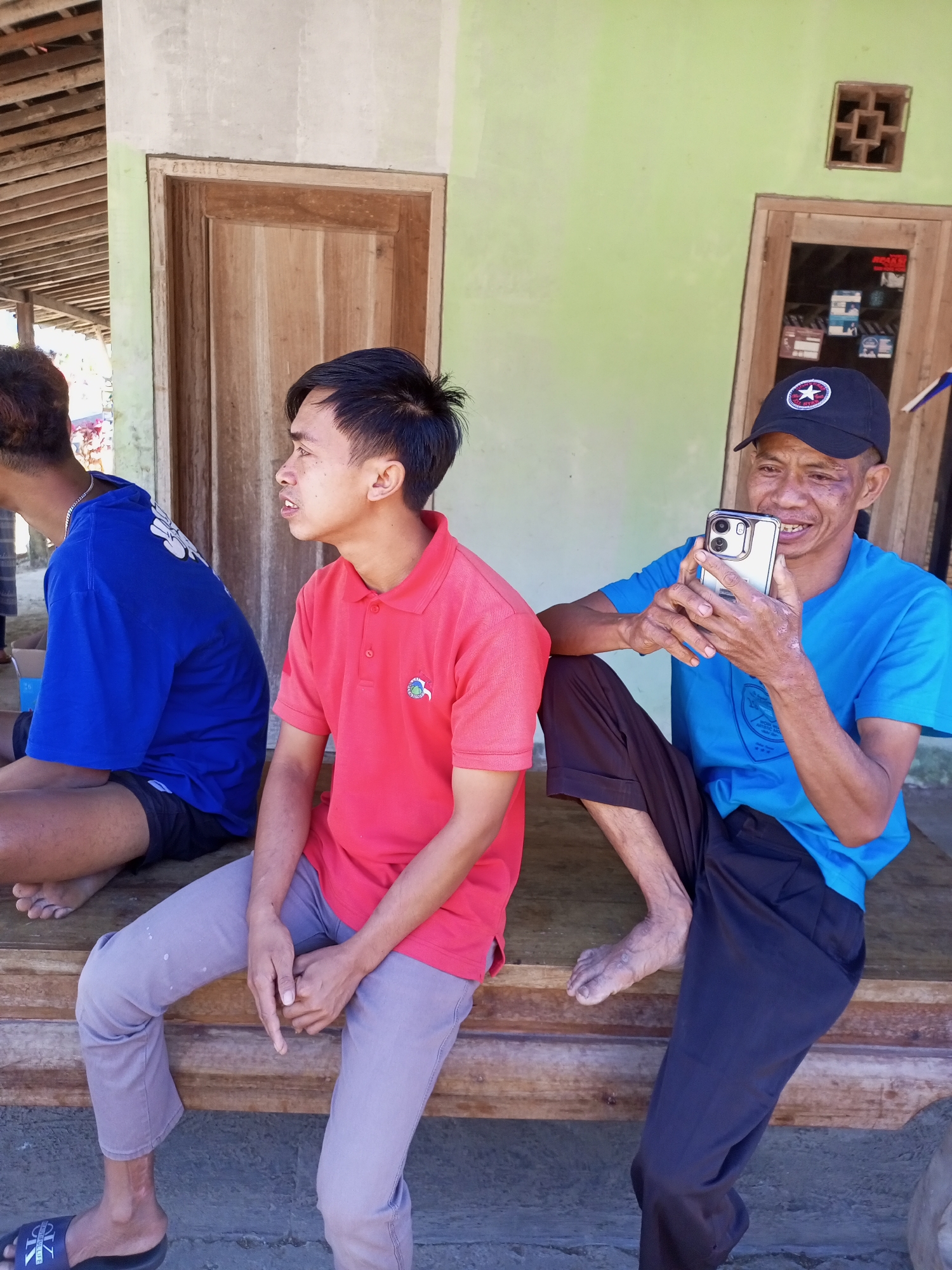 Three individuals are seated outdoors on a wooden structure in front of a building with light green walls and wooden doors. The person on the left is wearing a blue t-shirt and dark shorts. The person in the middle is wearing a red polo shirt and light gray pants. The person on the right is wearing a blue t-shirt, dark pants, and a black baseball cap. This individual is holding a silver smartphone in their hands and appears to be smiling while looking at the screen. The scene suggests a casual gathering or interaction in a rural or semi-rural setting, consistent with locations in Jatipurno, Indonesia, known for its traditional architecture and community spaces.