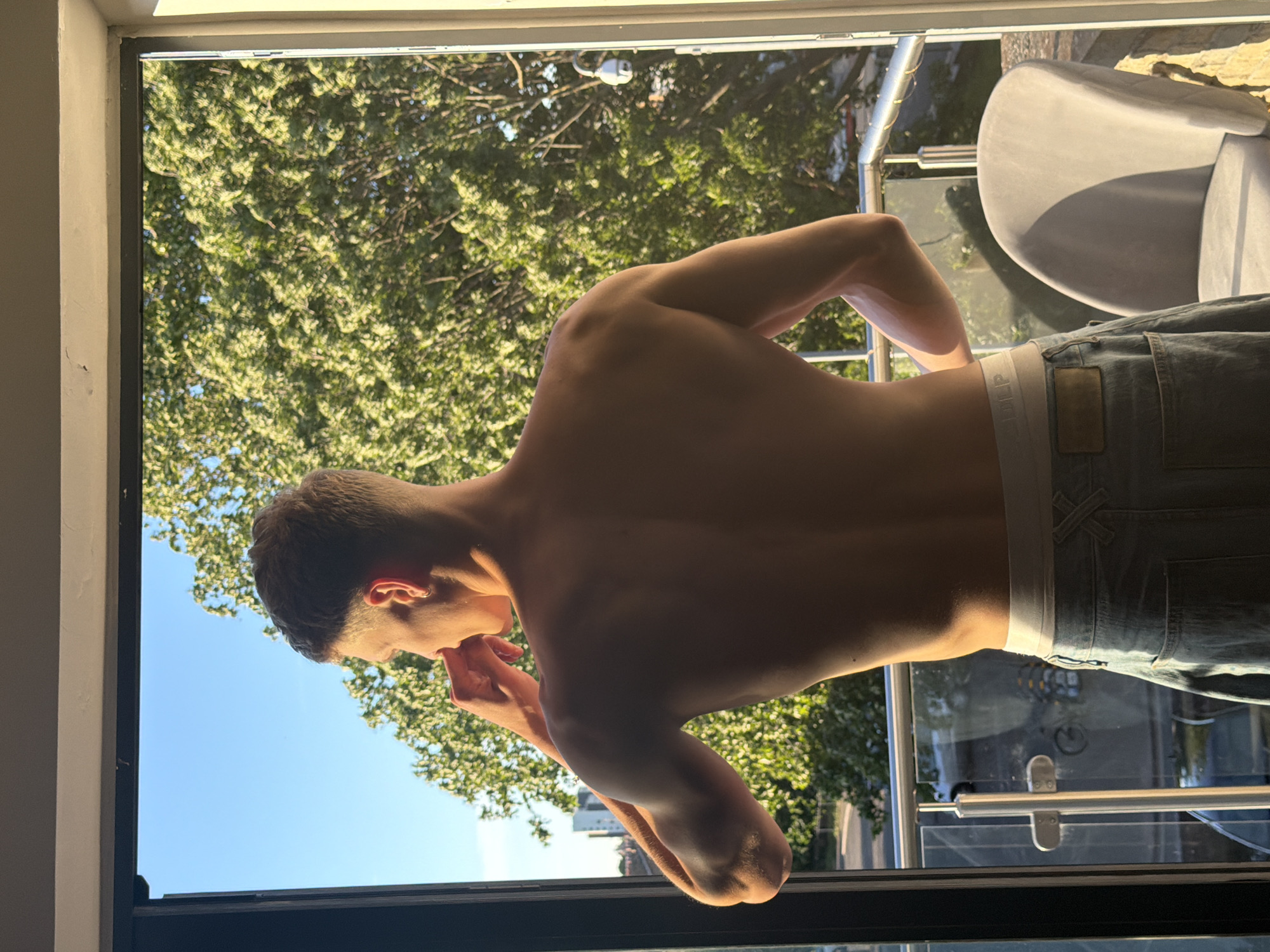A young, shirtless man with short brown hair stands on a balcony, looking away from the camera. He is wearing light blue jeans. Sunlight illuminates his back and arms, highlighting his muscular physique. In the background, lush green trees and a clear blue sky suggest a warm, sunny day. A grey chair is visible on the balcony. The scene appears to be in a residential area within the city of London, United Kingdom, as indicated by the presence of buildings and a street visible beyond the balcony railing.