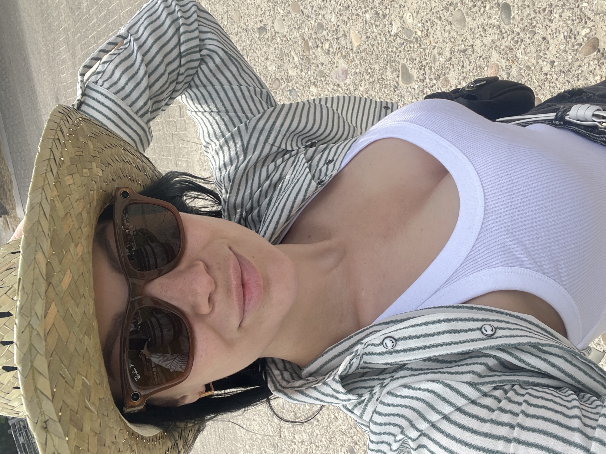 A woman, wearing a straw hat, sunglasses, and a striped button-up shirt over a white tank top, is taking a selfie outdoors. The background suggests an outdoor setting with gravel and a paved surface. The sunglasses reflect a scene with barrels and a person holding a phone, indicating the woman is taking the photo. The woman's hand is raised, adjusting her hat.