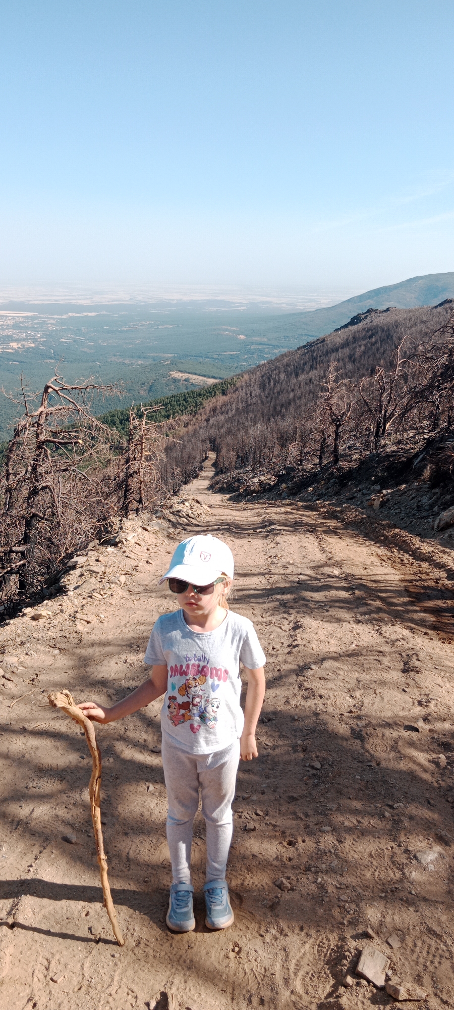 A young girl, wearing a white baseball cap, sunglasses, a t-shirt with a Paw Patrol graphic, grey sweatpants, and blue sneakers, stands on a dirt path. She is holding a stick in her left hand. The path appears to be in a mountainous area with evidence of a past fire, as many trees are bare and charred. The background shows a vast landscape with green forests and a distant hazy horizon, characteristic of the region around Cerezo de Arriba, Spain. The sky is clear and blue.