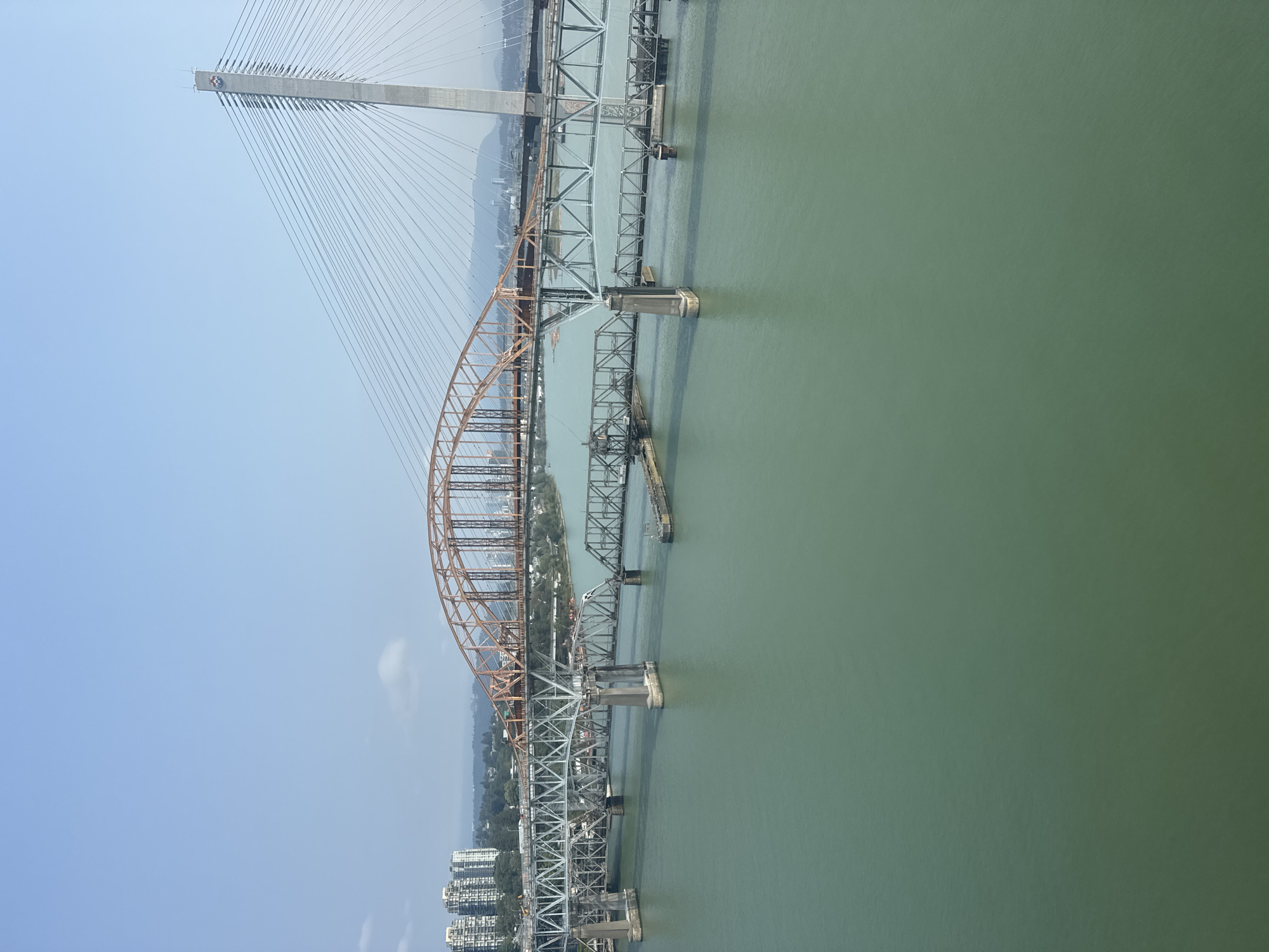 The image captures an aerial view of a multi-bridge crossing over a body of water, likely a river or inlet, in Surrey, Canada. The prominent structures are three distinct bridges positioned parallel to each other. The foremost bridge, visible from the right side of the frame, is a modern cable-stayed bridge with a tall concrete pylon from which numerous cables extend to support the roadway. To its left is a large, orange steel arch bridge, also a significant structure. Further to the left, a lower, older-looking truss bridge with visible riveted metalwork spans the water. This bridge appears to be supported by multiple concrete piers. The water itself is a murky green color, with some small floating debris visible. In the background, the cityscape of Surrey is discernible, with high-rise residential buildings on the left bank and a more spread-out urban landscape beyond the bridges. Hills or mountains form the distant horizon under a clear, pale blue sky with a few wispy clouds. There are no people or vehicles clearly visible in the image, focusing the attention solely on the impressive engineering of the bridge system.