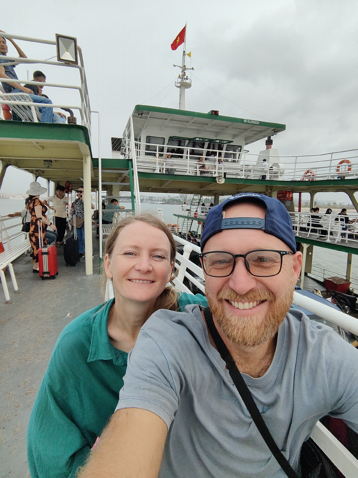A selfie photograph shows a man and a woman smiling on a multi-level boat on a cloudy day. The man is in the foreground, wearing a blue baseball cap, glasses, and a light gray t-shirt with a black strap over his shoulder. He has a red beard and is smiling broadly. The woman, slightly behind him, is wearing a green shirt and smiling.
In the background, other passengers can be seen on the boat, some with luggage. The boat has white railings and green trim, with a Vietnamese flag on the top level. The boat's cabin has the alphanumeric string "VR 04010231." The scene seems to be Tuan Chau Ward, Vietnam, implied by the local architecture and flag. The image has a candid and casual feel.